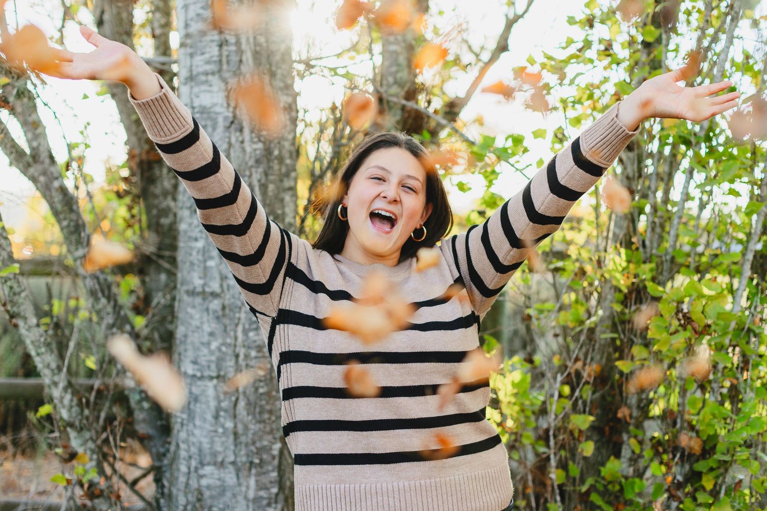 A joyful person in a striped sweater celebrates autumn by tossing fallen orange leaves in a sunlit forest.