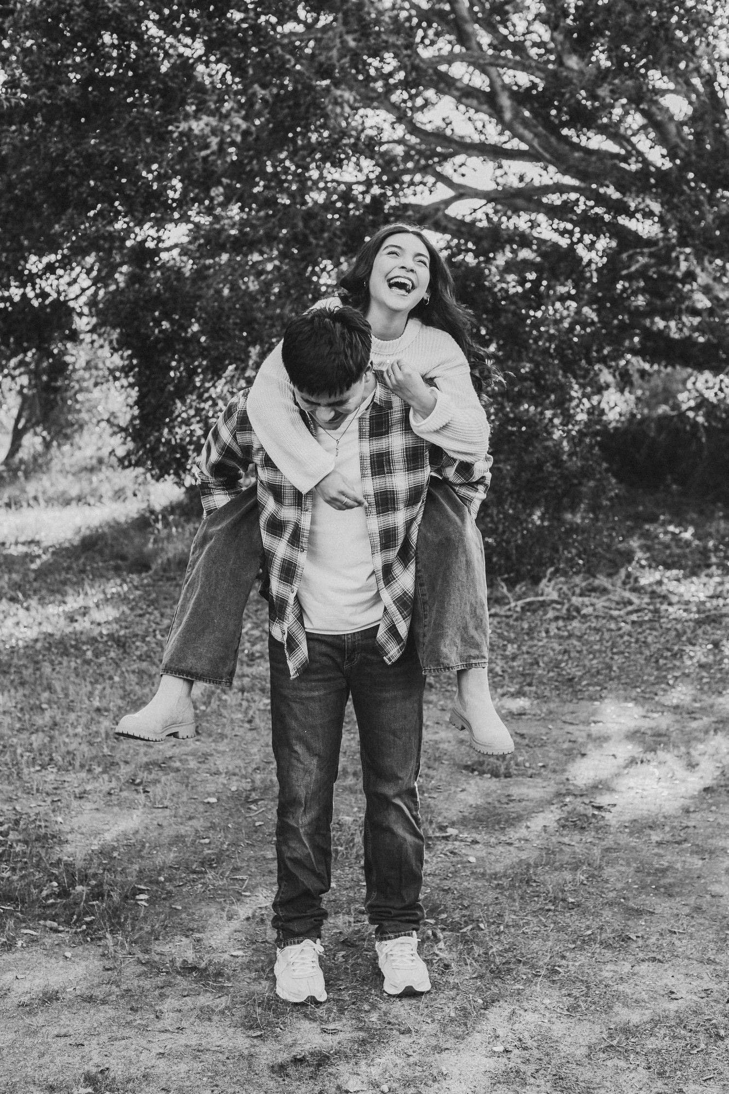 Black and white photo of a playful couple showing joyful expressions outdoors with trees in background.