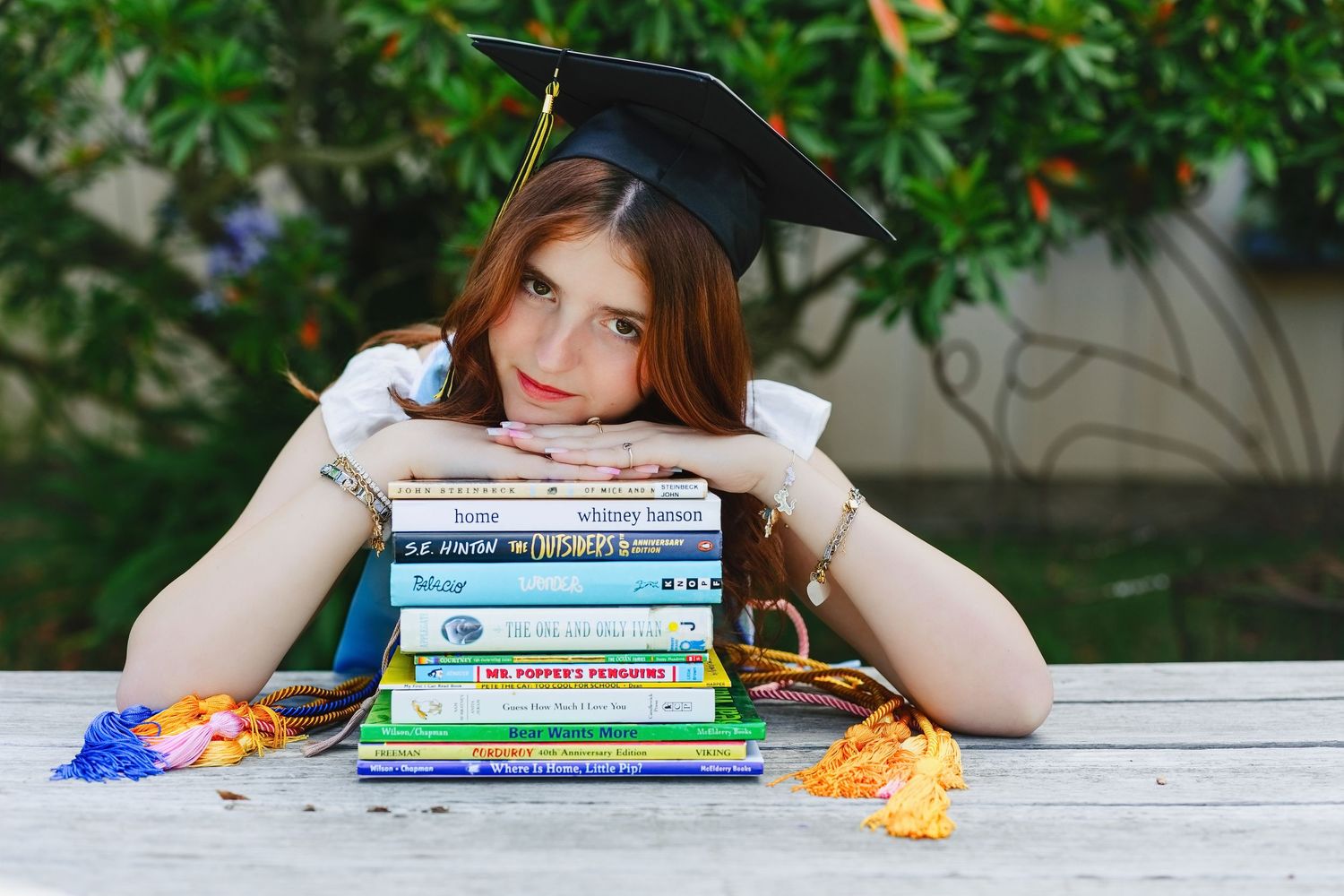 Young woman in graduation cap resting on stack of colorful books at outdoor wooden table.