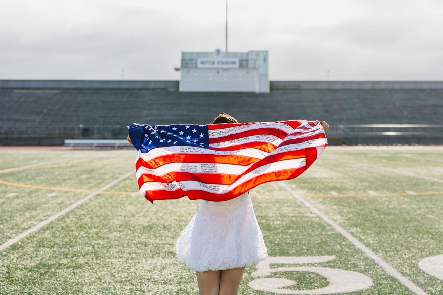 Young woman in white dress holding an American flag on a football field stadium.