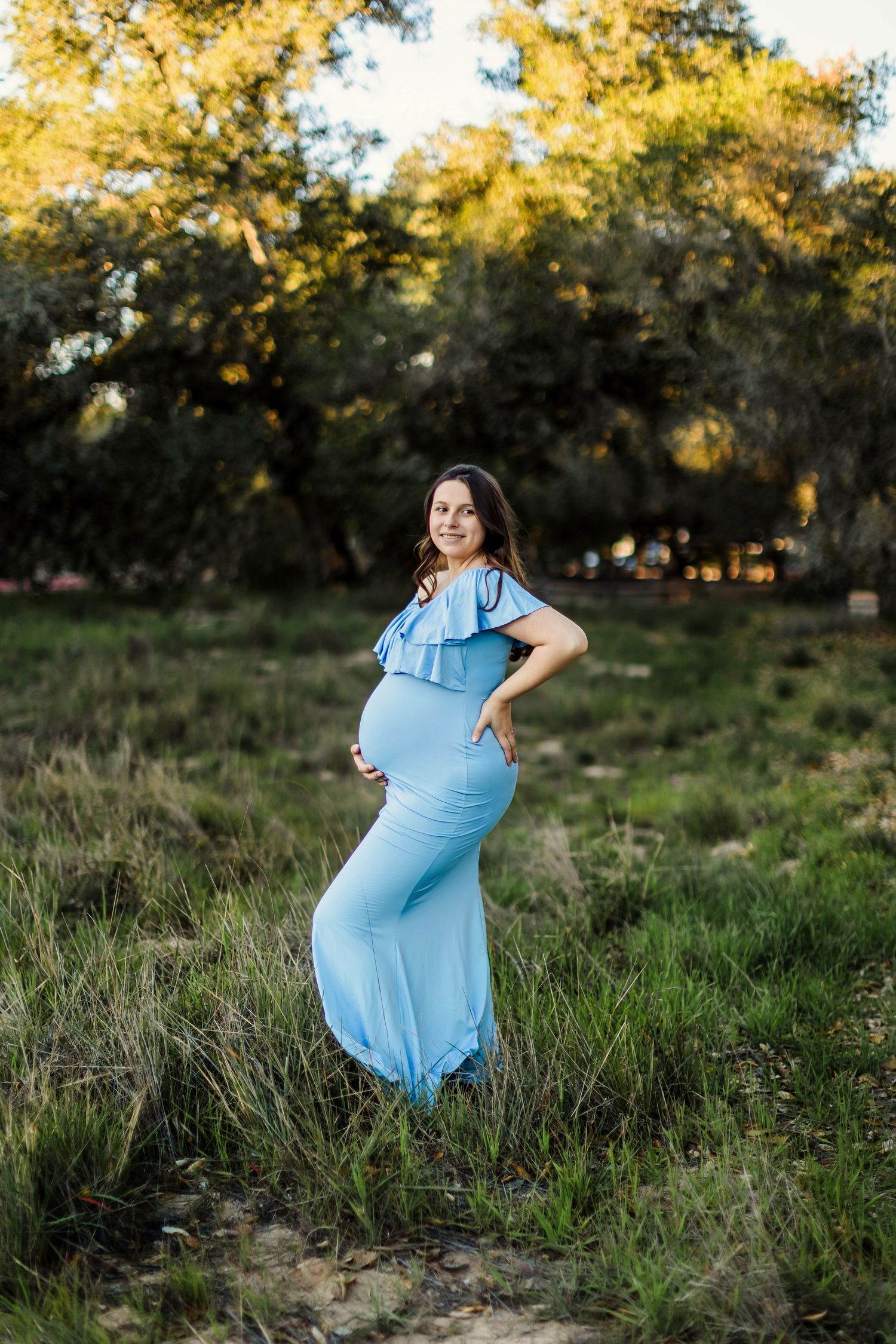 A pregnant woman in a flowing light blue dress poses gracefully in a sunlit meadow during golden hour.