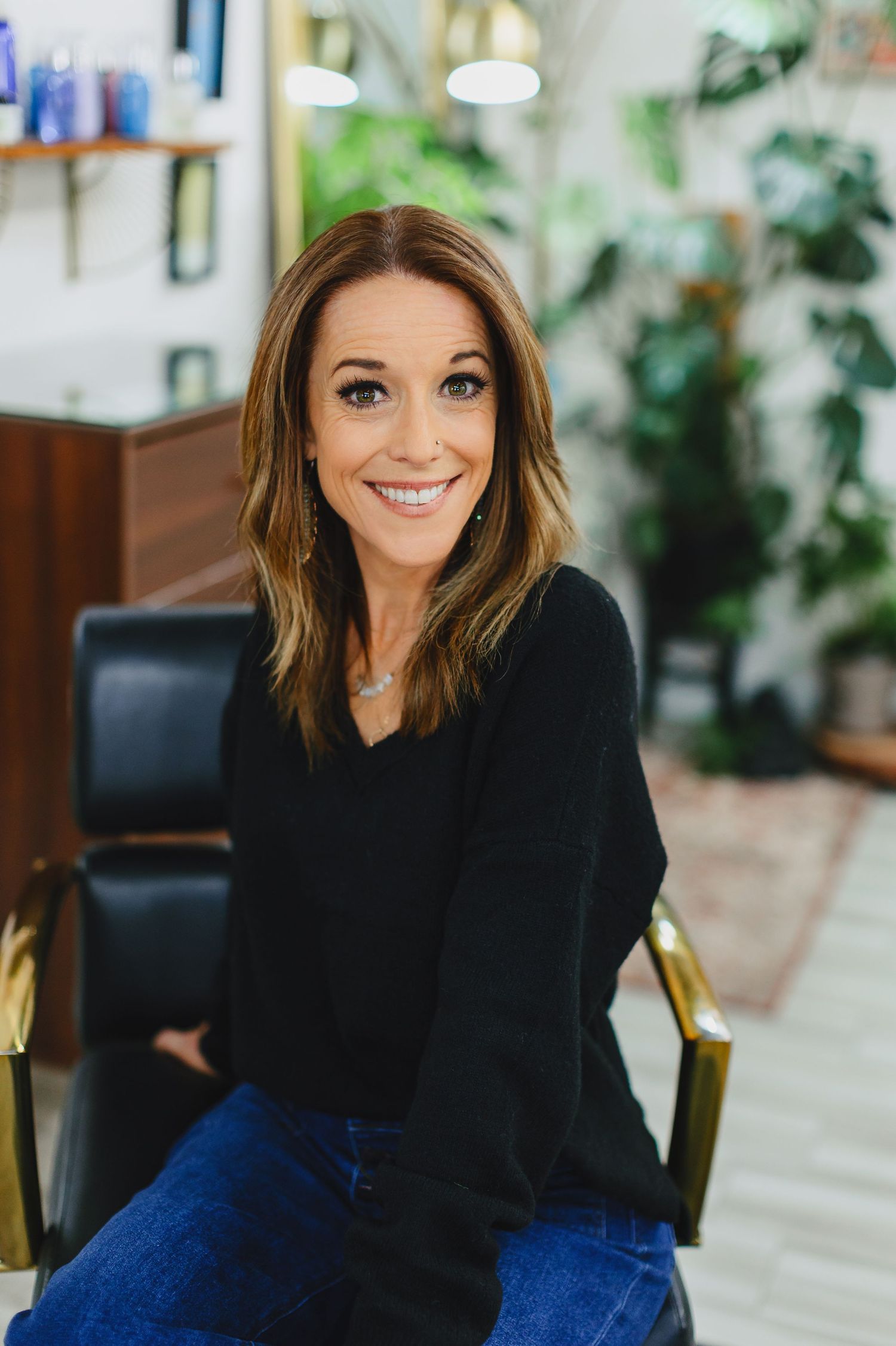 Professional businesswoman in black sweater and jeans sitting casually in office with plants in background.