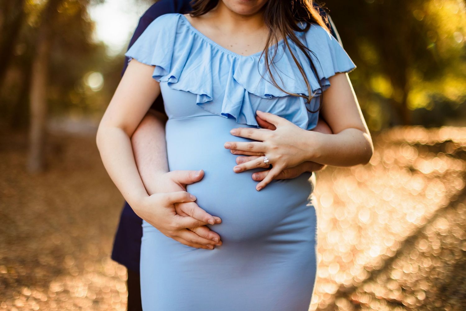 Pregnant woman in soft blue dress holds her belly during golden hour maternity photoshoot in nature.
