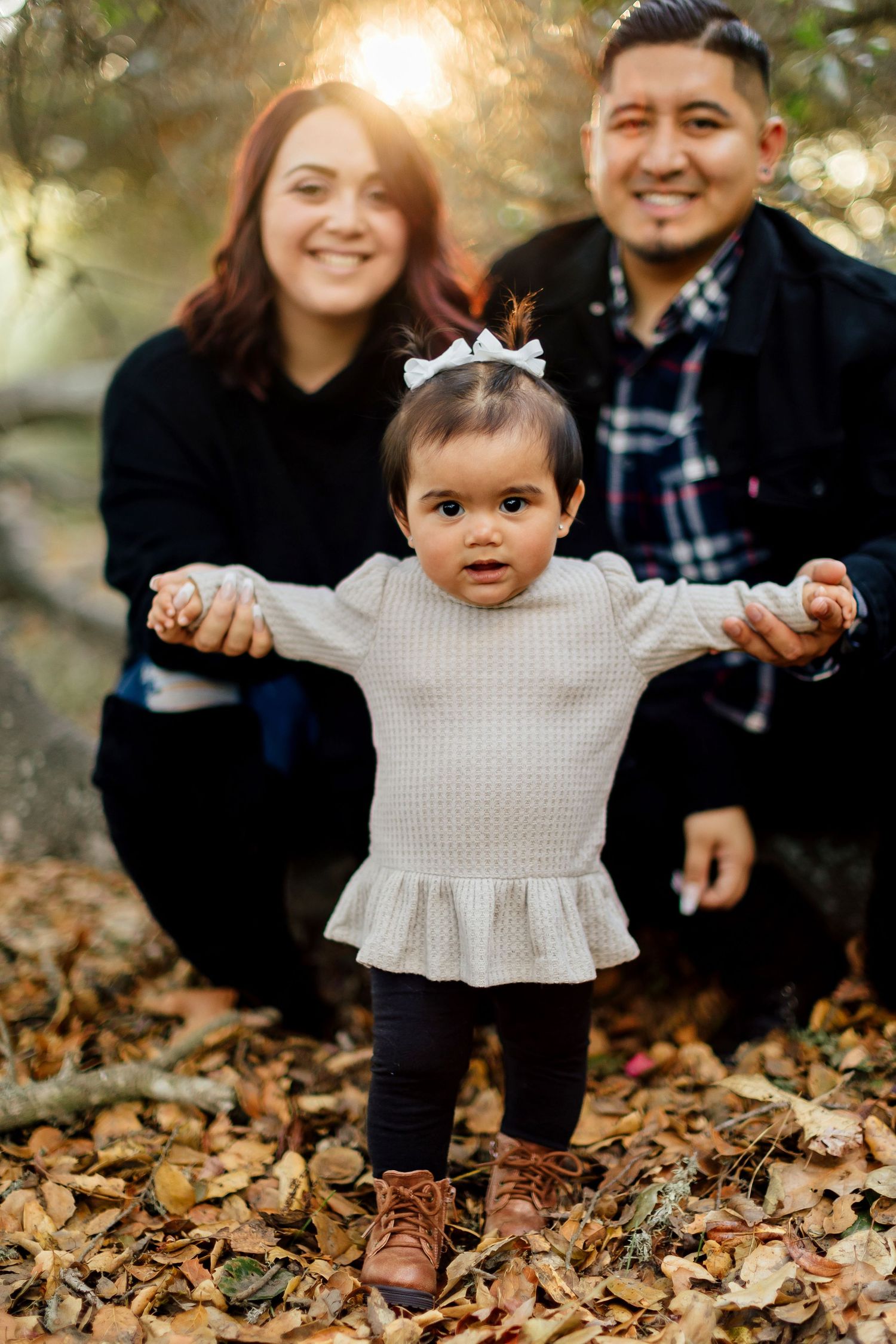 An adorable toddler in a ruffled sweater stands on autumn leaves while being photographed with family outdoors.