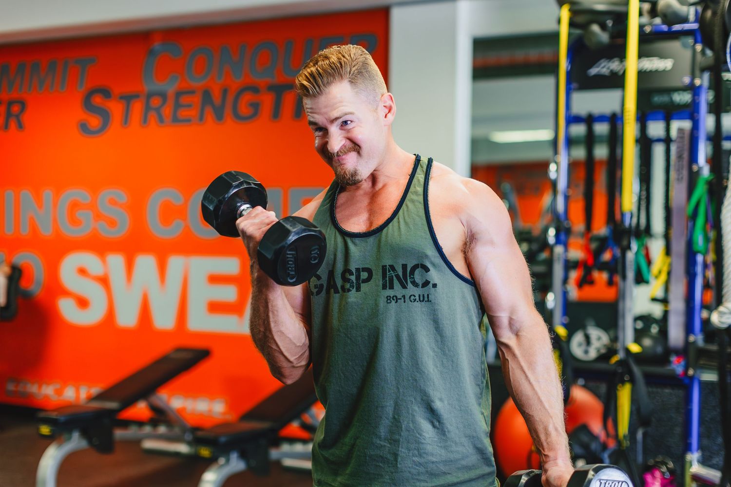 A muscular person performs bicep curls with a dumbbell in a vibrant orange and blue gym setting.