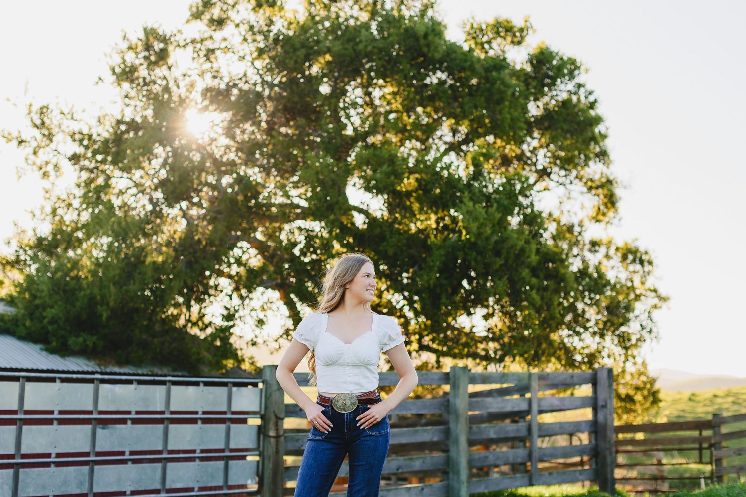 Blonde woman in white top and jeans posing by a wooden fence with sunlight through trees.