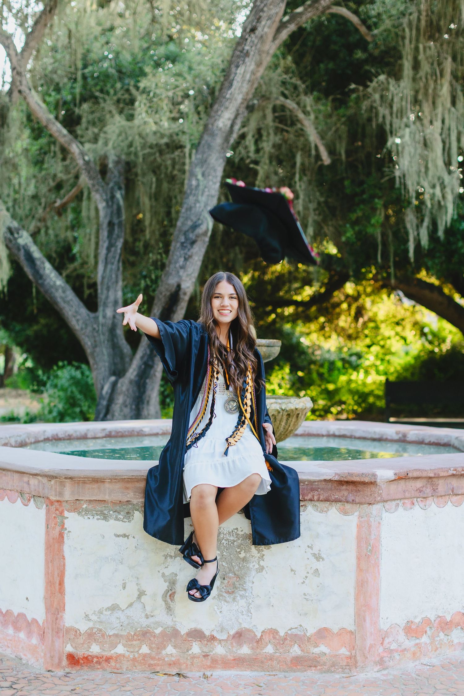 Smiling graduate in black cap and gown sitting by a fountain tossing her cap in a garden.