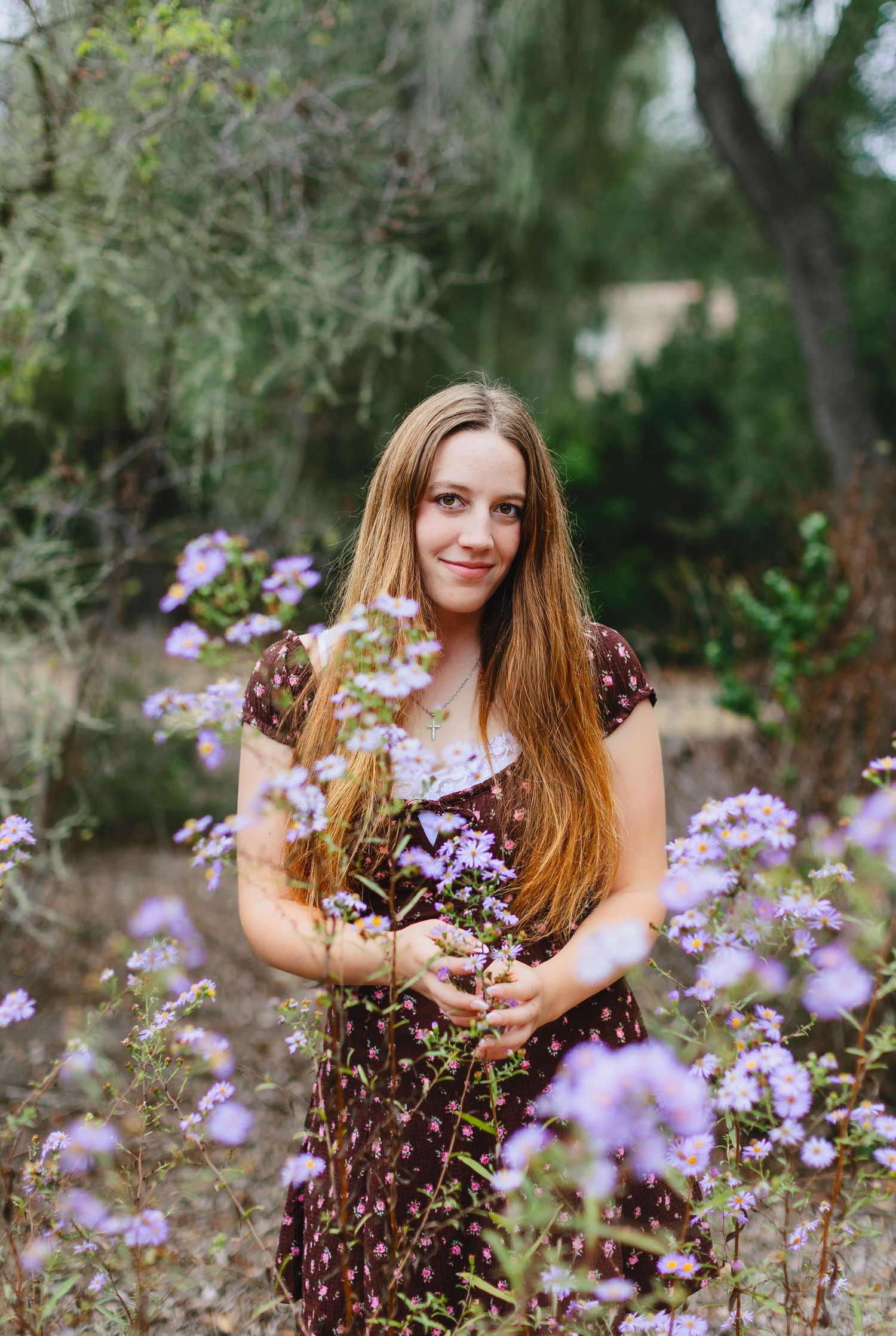 Young woman with long hair in floral dress holding purple wildflowers in a lush garden setting.