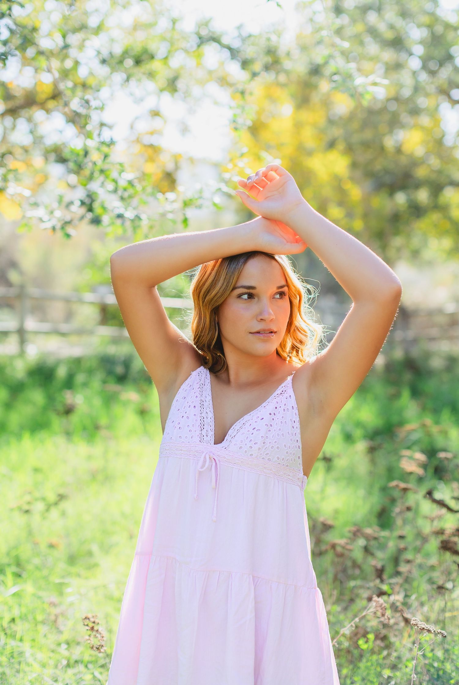 Young woman in pink sundress poses with arms raised outdoors in a sunny green garden setting.