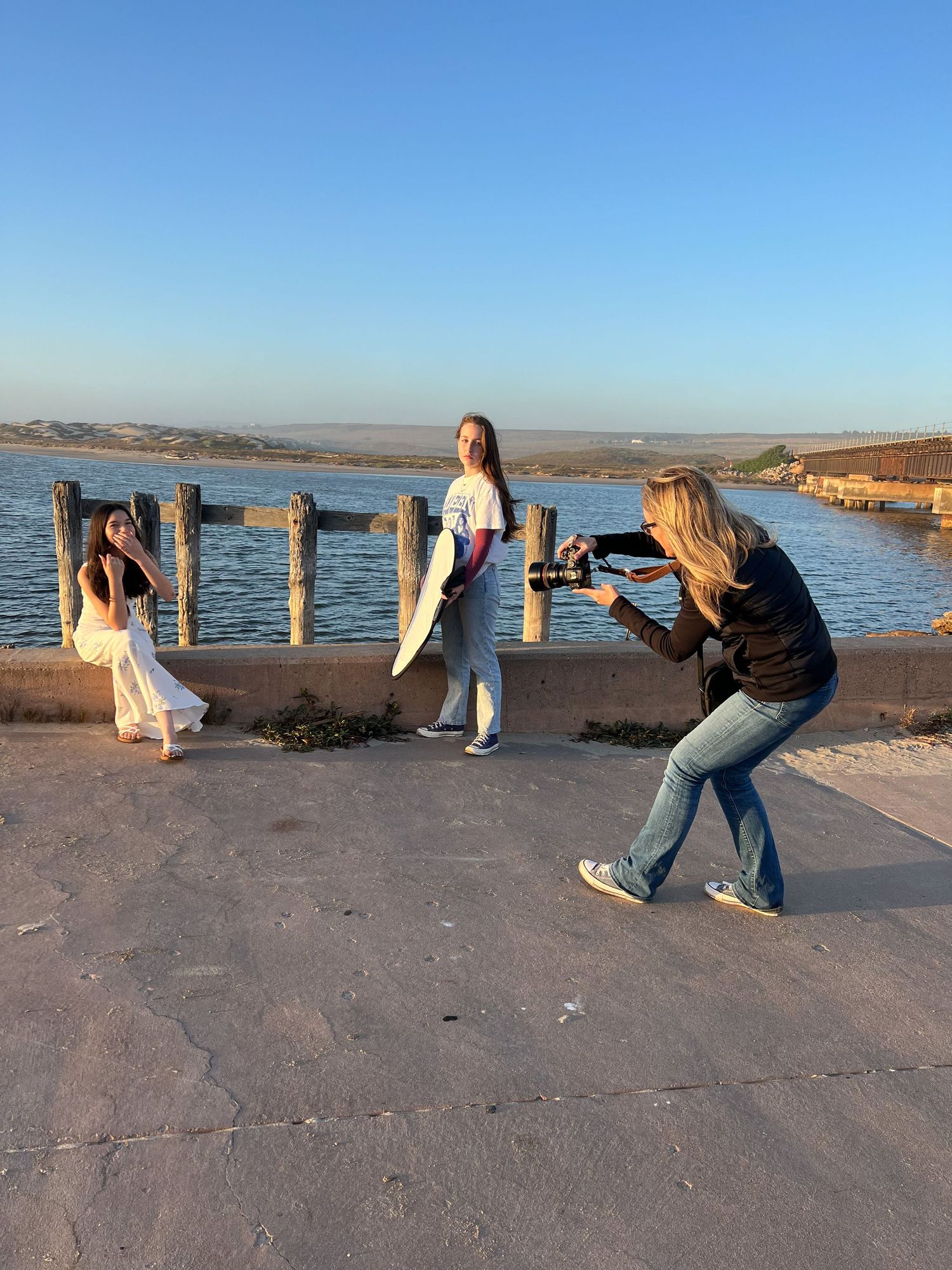 Photographer shoots two girls posing near waterfront dock during golden hour sunset by the bay.