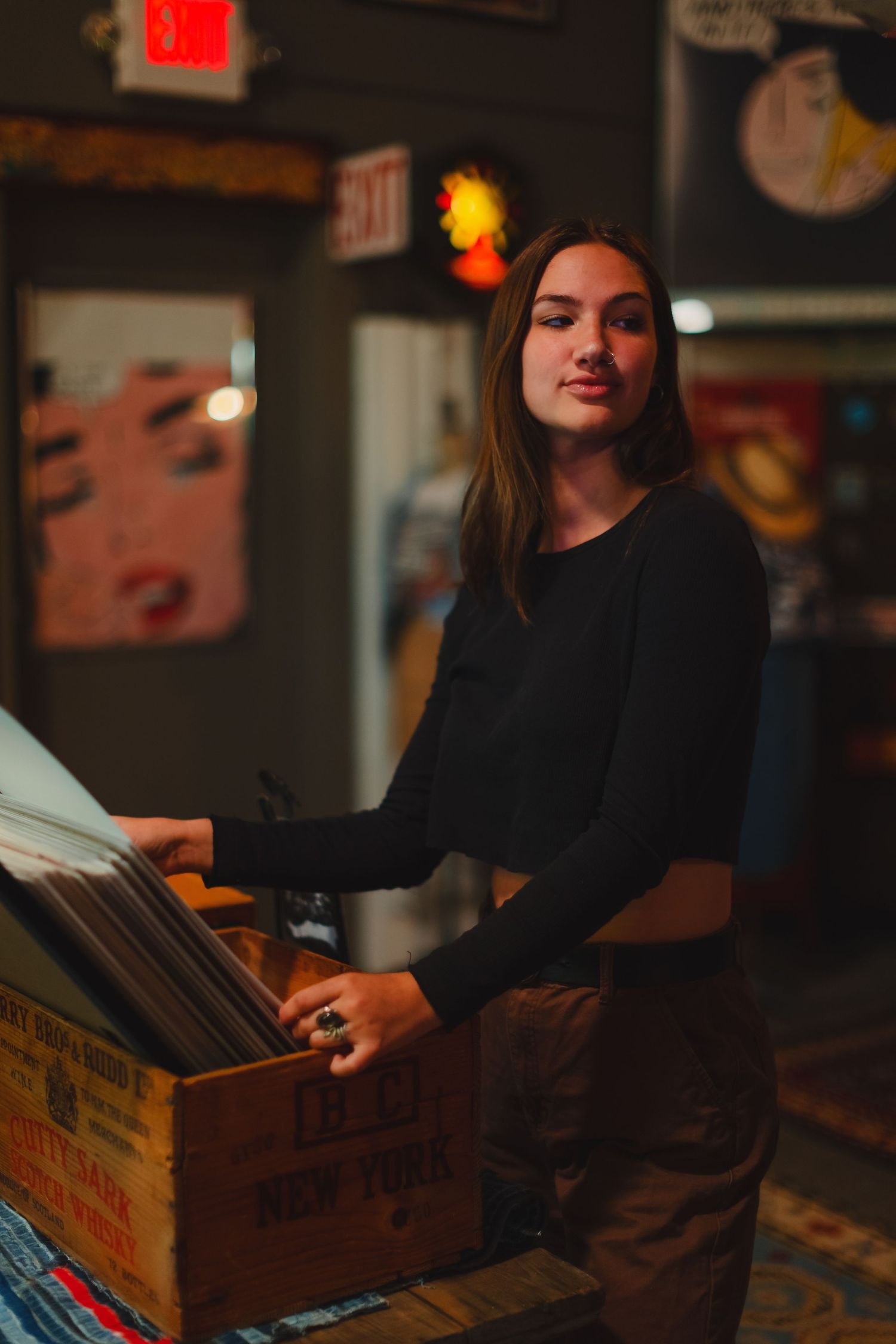 Person browsing through vinyl records in a wooden crate at a dimly lit record store.