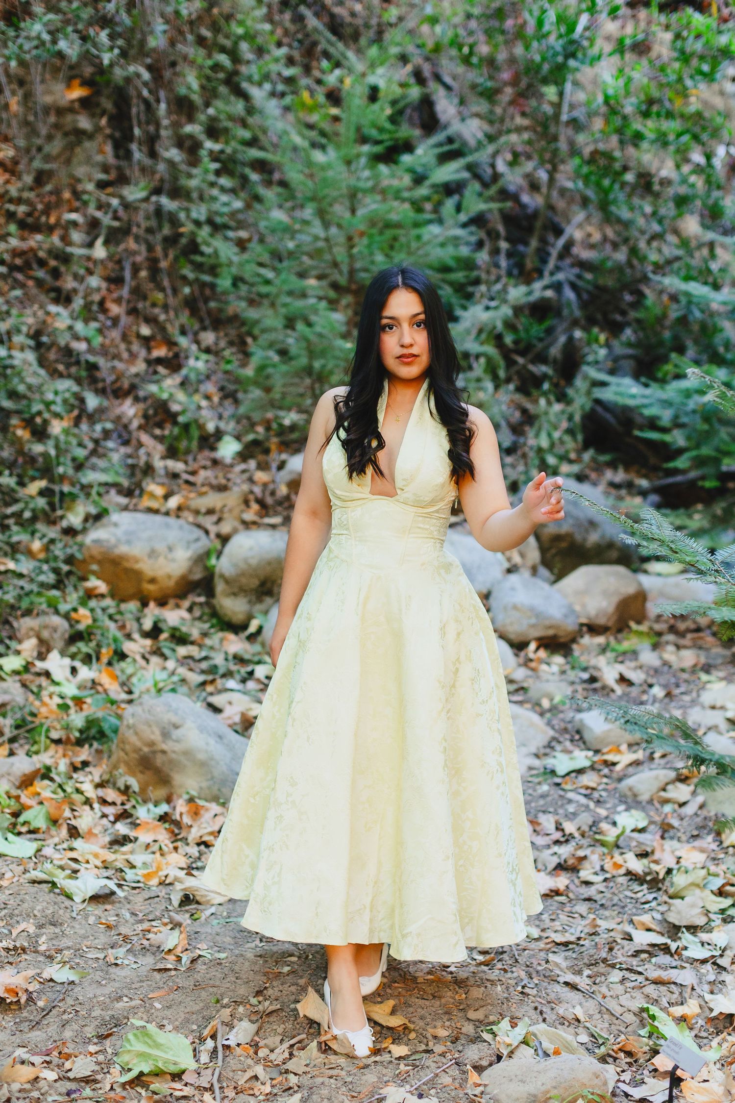 Young woman in elegant yellow vintage halter dress standing on a rocky forest trail.