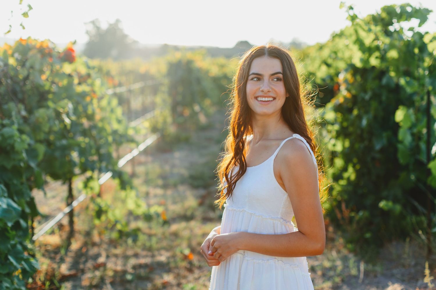 Smiling young woman in white sundress stands in sunny vineyard with green vines behind her.
