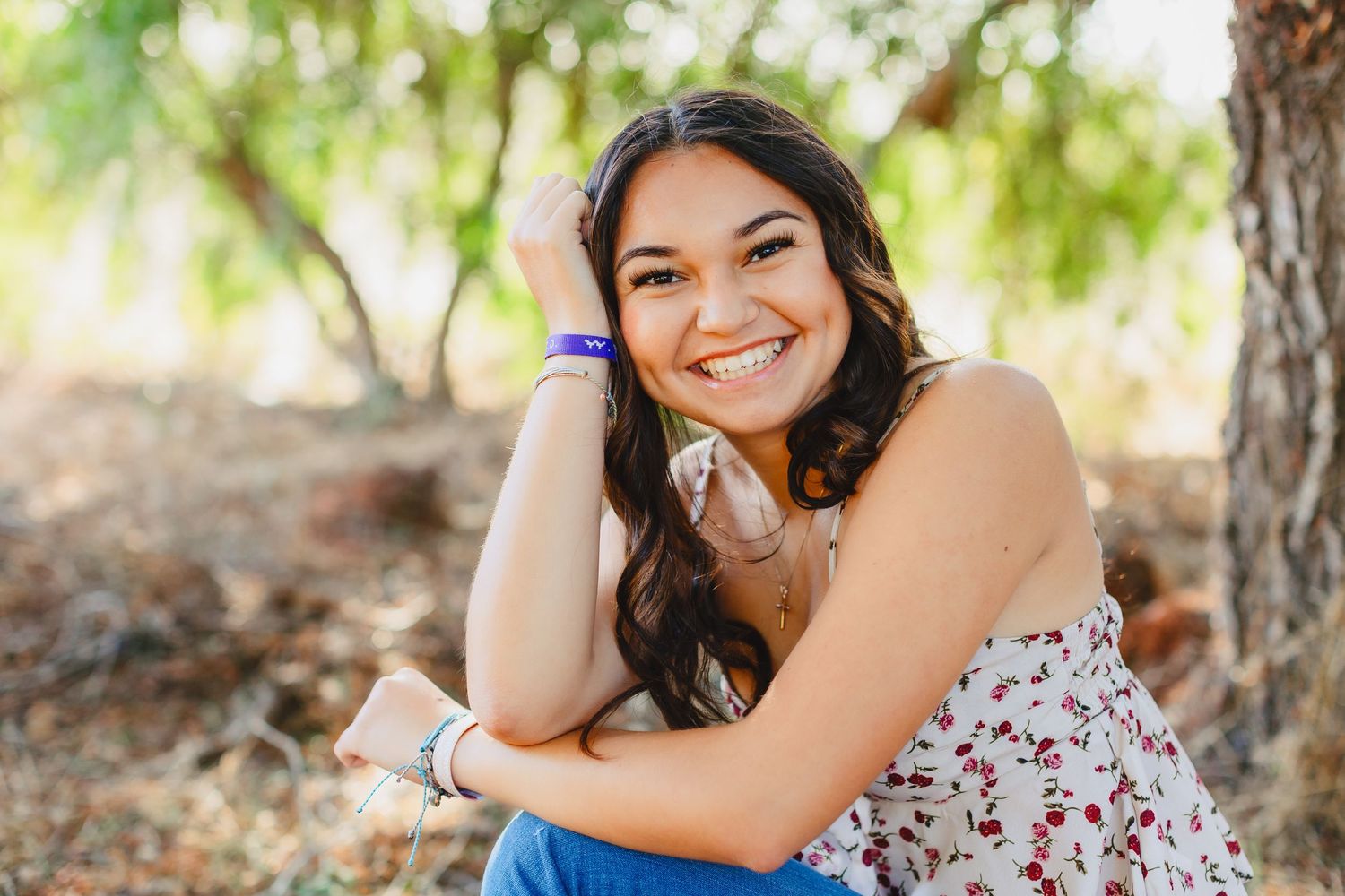 Smiling young woman in floral top and blue jeans sitting outdoors in natural lighting with trees in background.