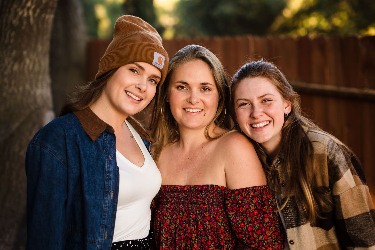 Three friends share a warm smile outdoors while posing together in casual fall attire for a portrait.