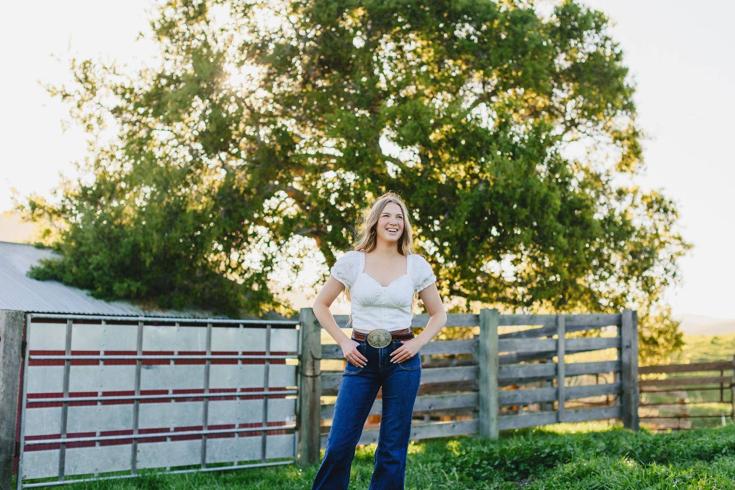 Woman in white top poses against rustic fence and tree backdrop during golden hour.