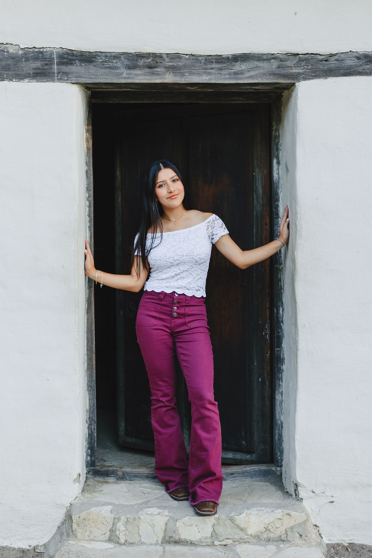 Young woman in white lace top and magenta flare jeans posing in a rustic wooden doorway of a white stucco wall.