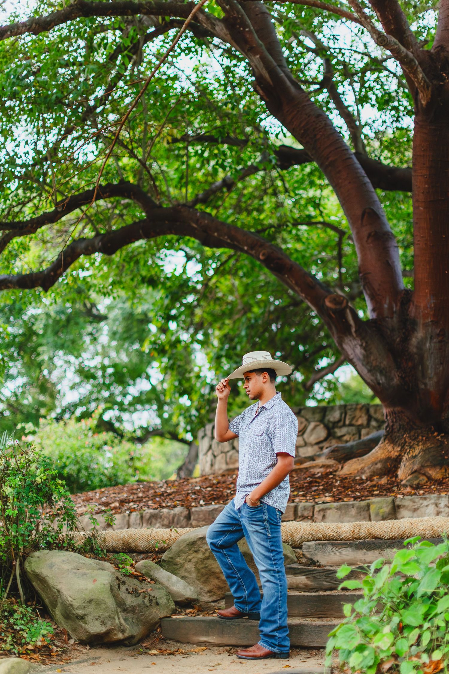 Young man in cowboy hat and jeans standing on stone steps beneath large oak trees.