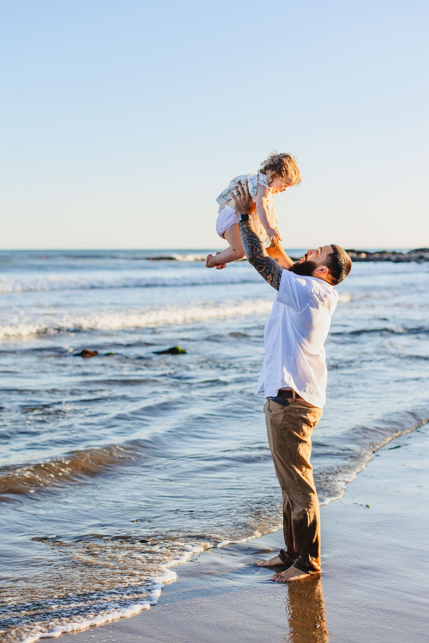 A parent lifts a child playfully in the air at the beach during sunset.