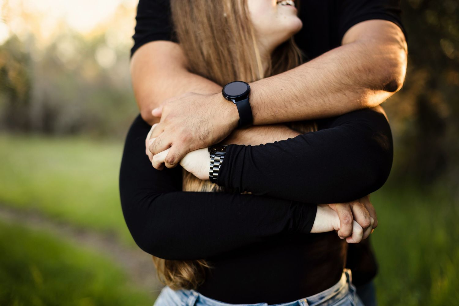 Close up of two people embracing outdoors in warm sunset light wearing black and denim clothing with a smartwatch visible.