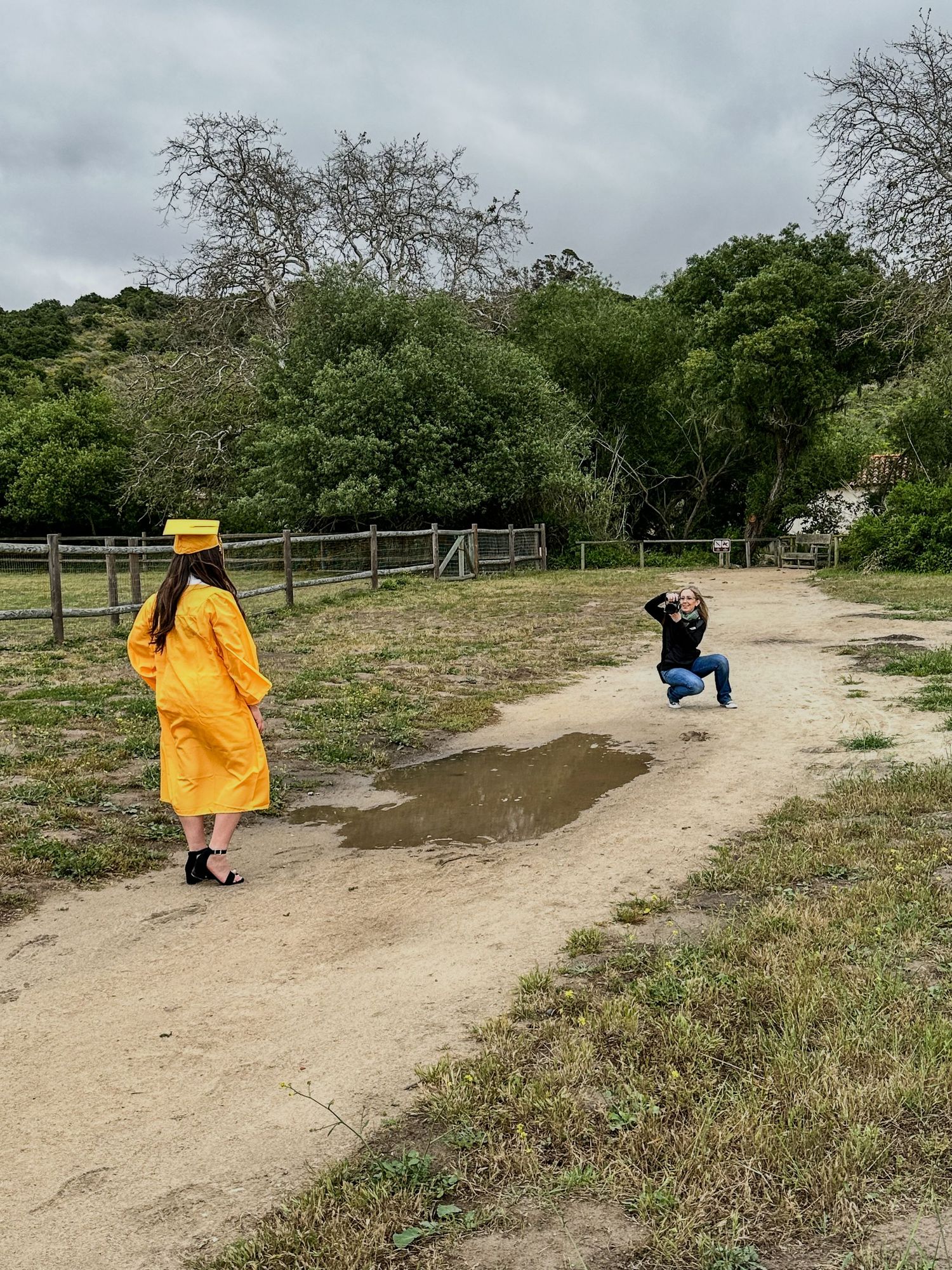 Photographer kneeling on dirt path photographing woman in yellow graduation cap and gown outdoors.