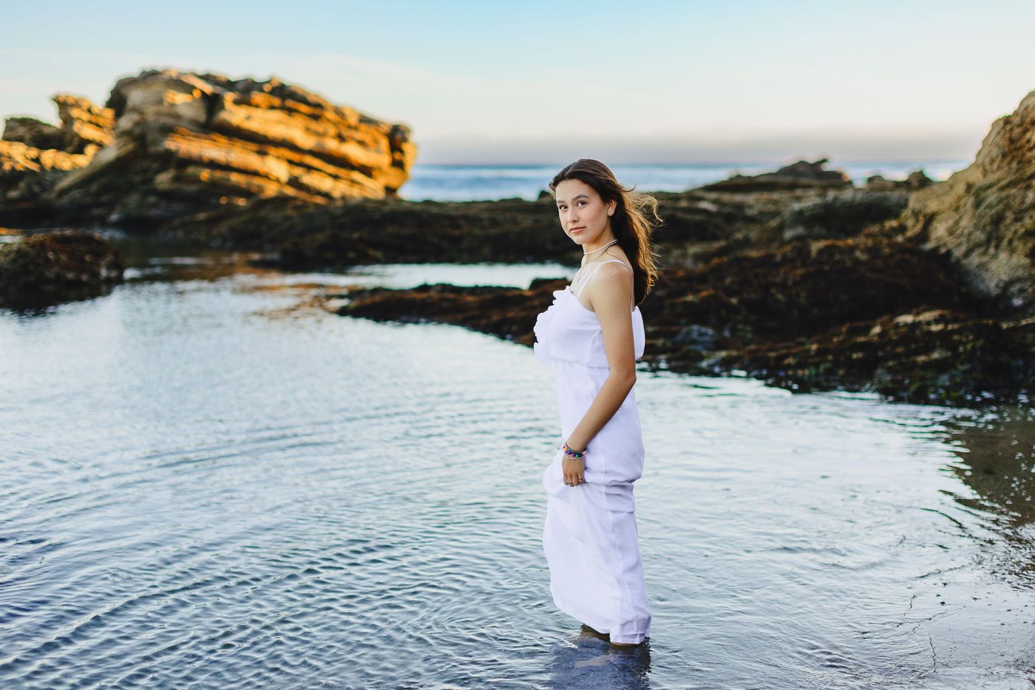 Person wearing white dress stands in shallow coastal waters at sunset with rocky cliffs in background.