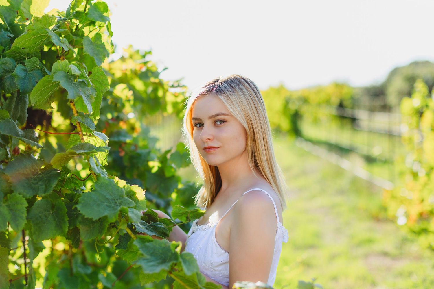A person in a white top poses in a sunlit vineyard with rows of green grapevines stretching into the distance.