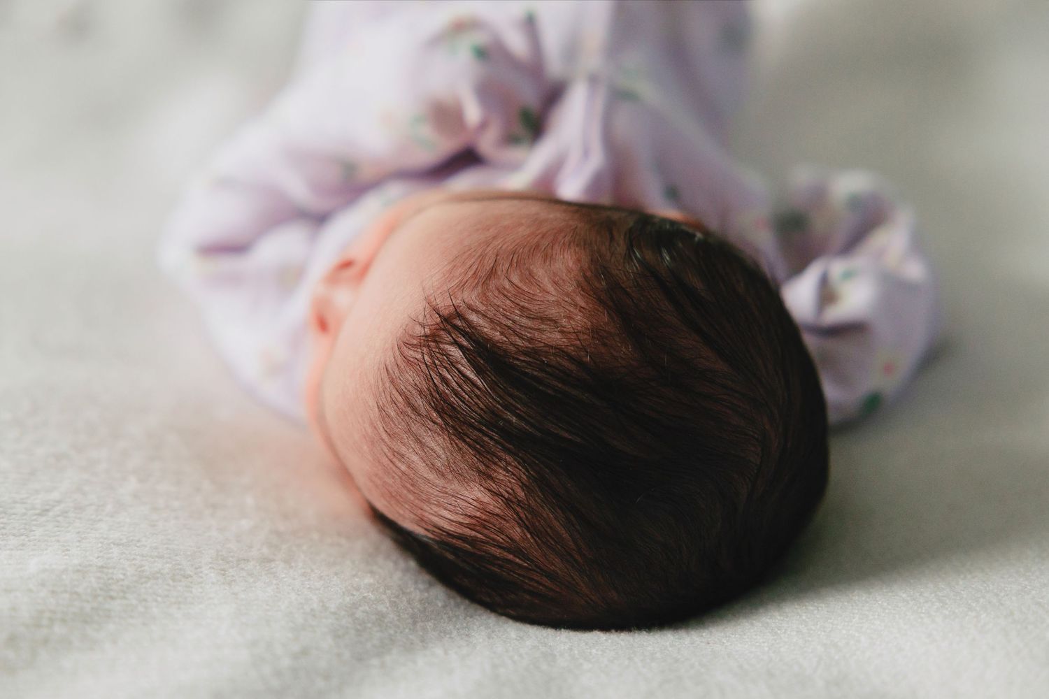 Newborn baby sleeping peacefully with dark hair visible, dressed in a soft lavender floral outfit on a light blanket.