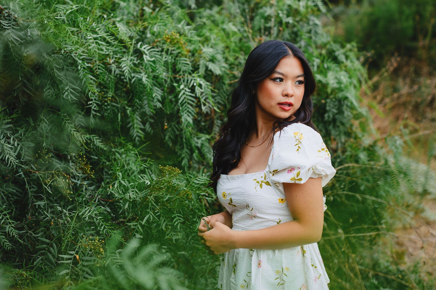 A woman in a white summer dress poses against lush green foliage outdoors during a sunny day.