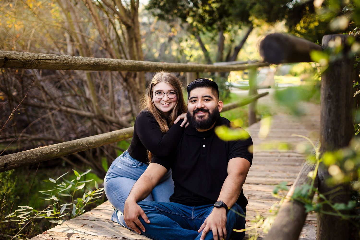 A couple sits close together on a wooden bridge surrounded by lush greenery on a sunny afternoon.