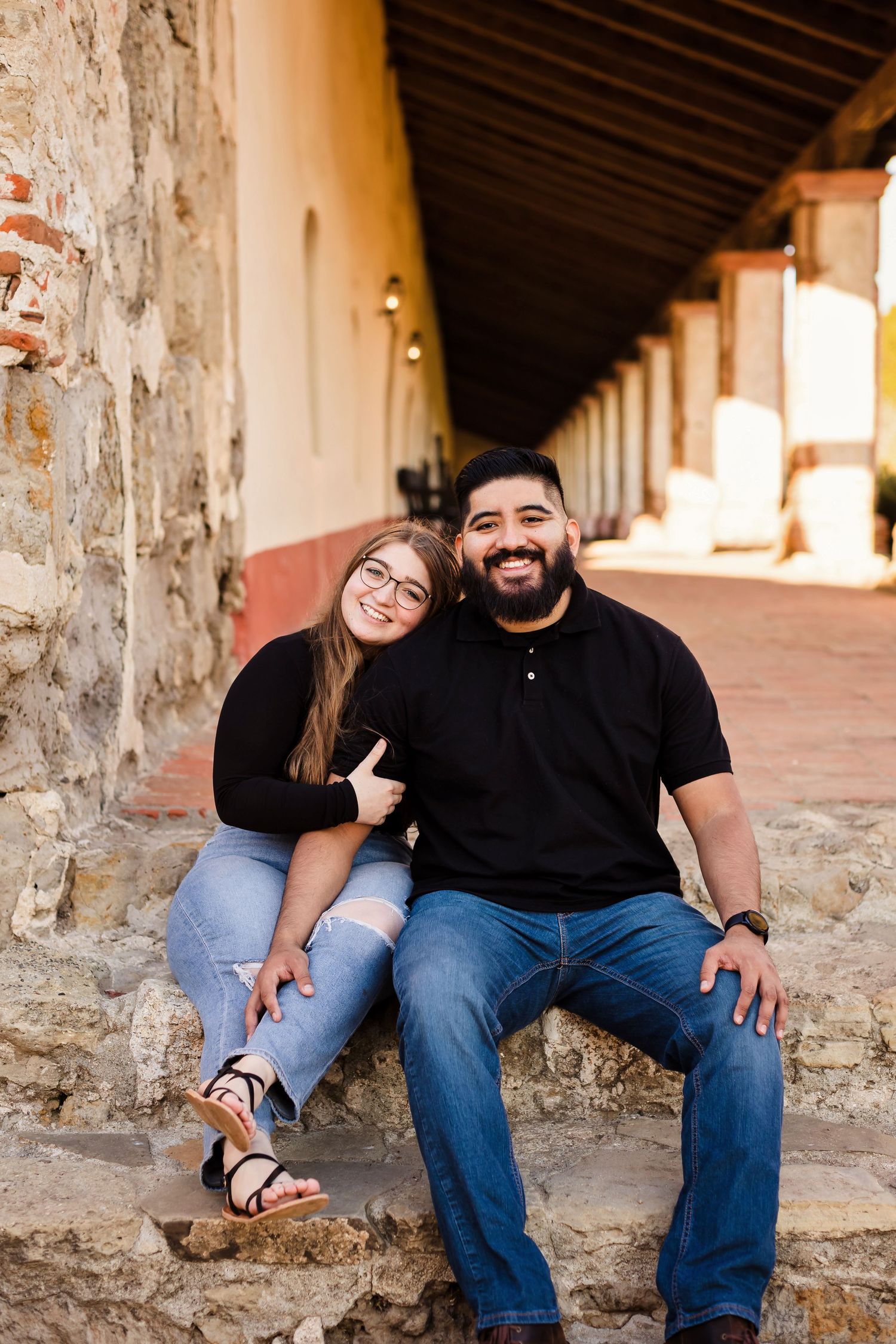 A couple sits together on stone steps in a rustic outdoor corridor with warm lighting and architectural details.