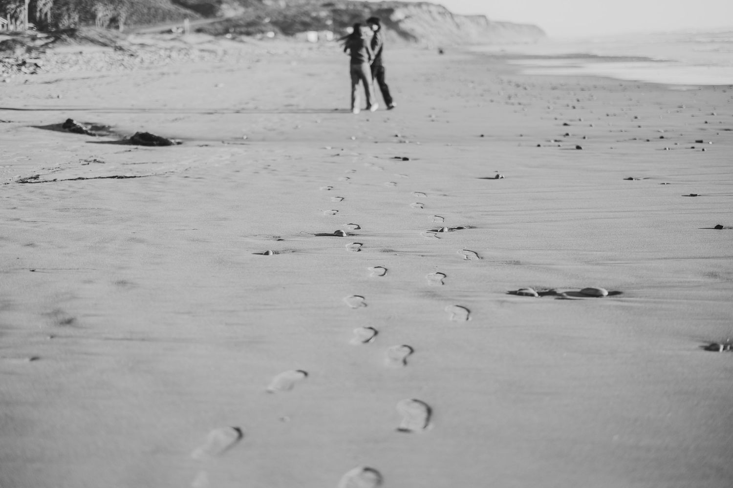 Black and white photo of a couple standing on a sandy beach with footprints leading toward them.