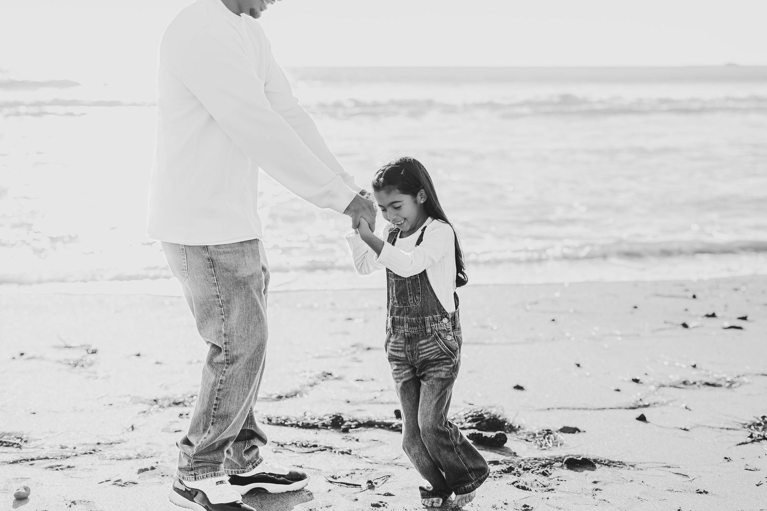 Black and white photo of a parent and child holding hands on a beach, child wearing overalls.