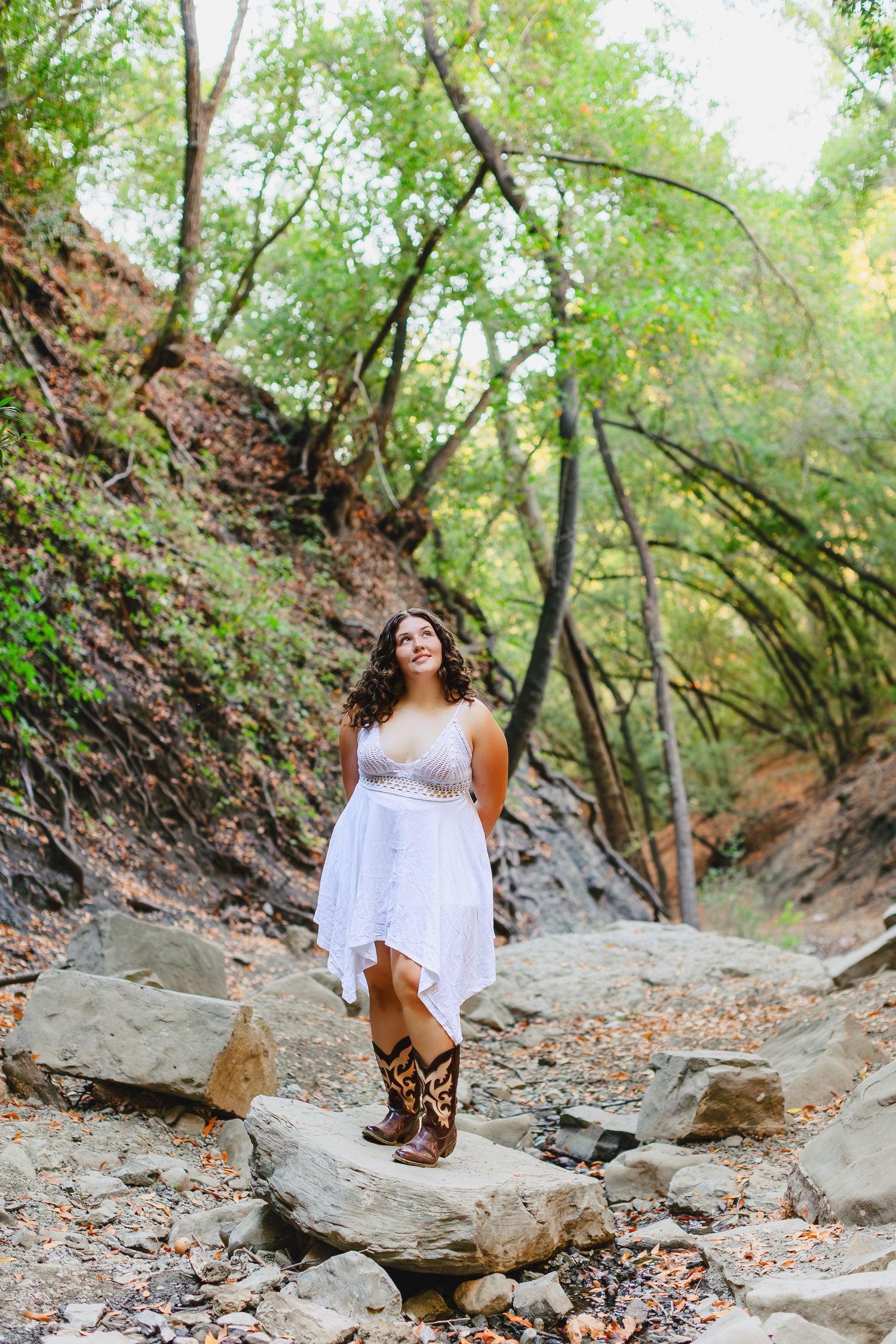 Young woman in white dress and cowboy boots standing on rocks in a lush green forest setting.