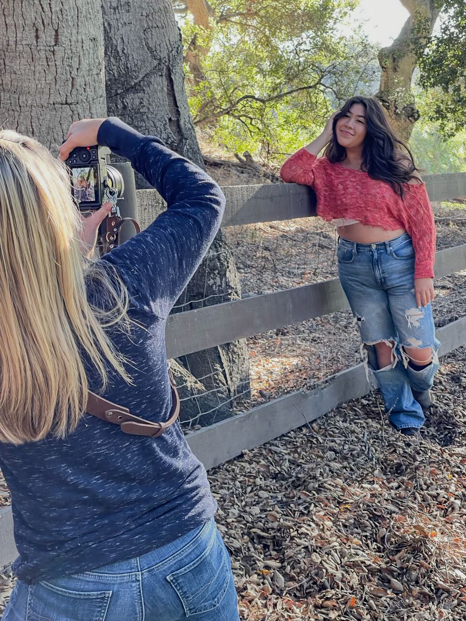 Blonde woman photographing a young brunette woman posing by a wooden fence outdoors.