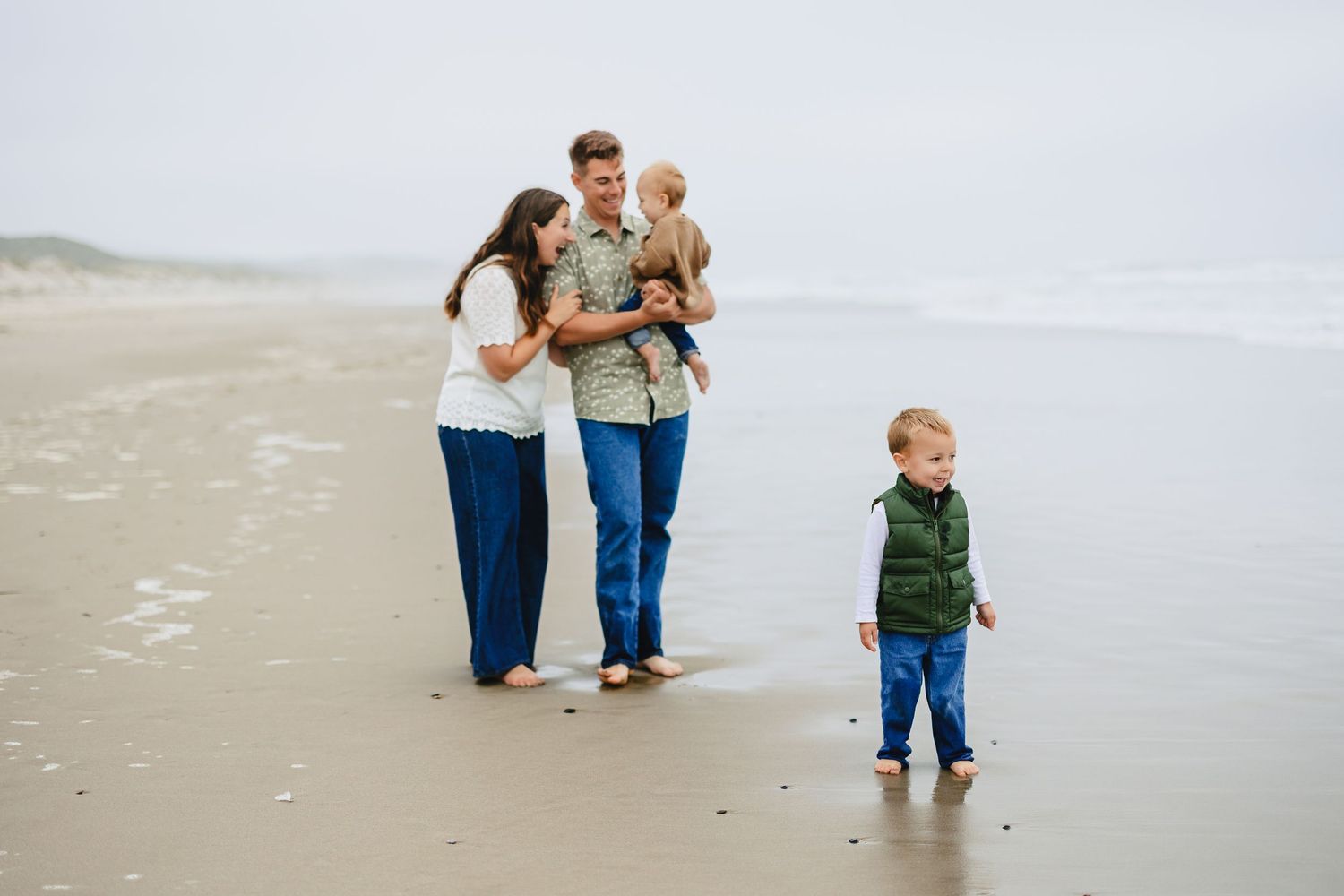 Family of four enjoying a beach walk on a cloudy day, parents holding toddler while young boy explores the shore.