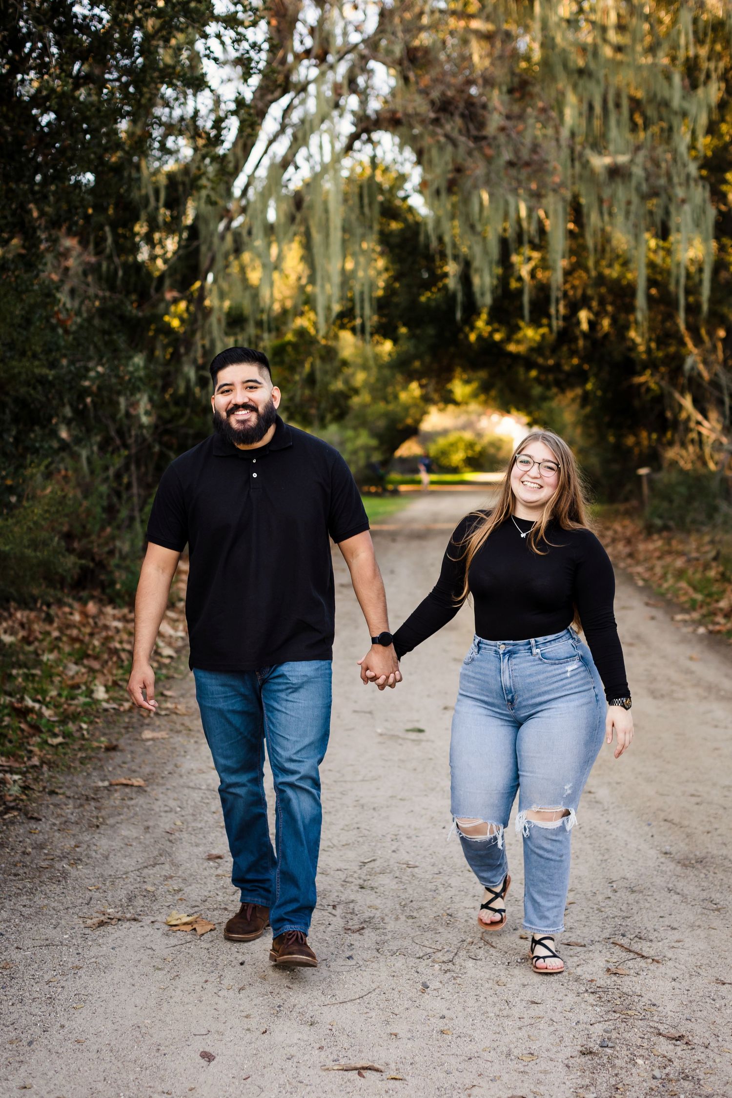 Two people walk together holding hands on a dirt path through Spanish moss-draped trees.