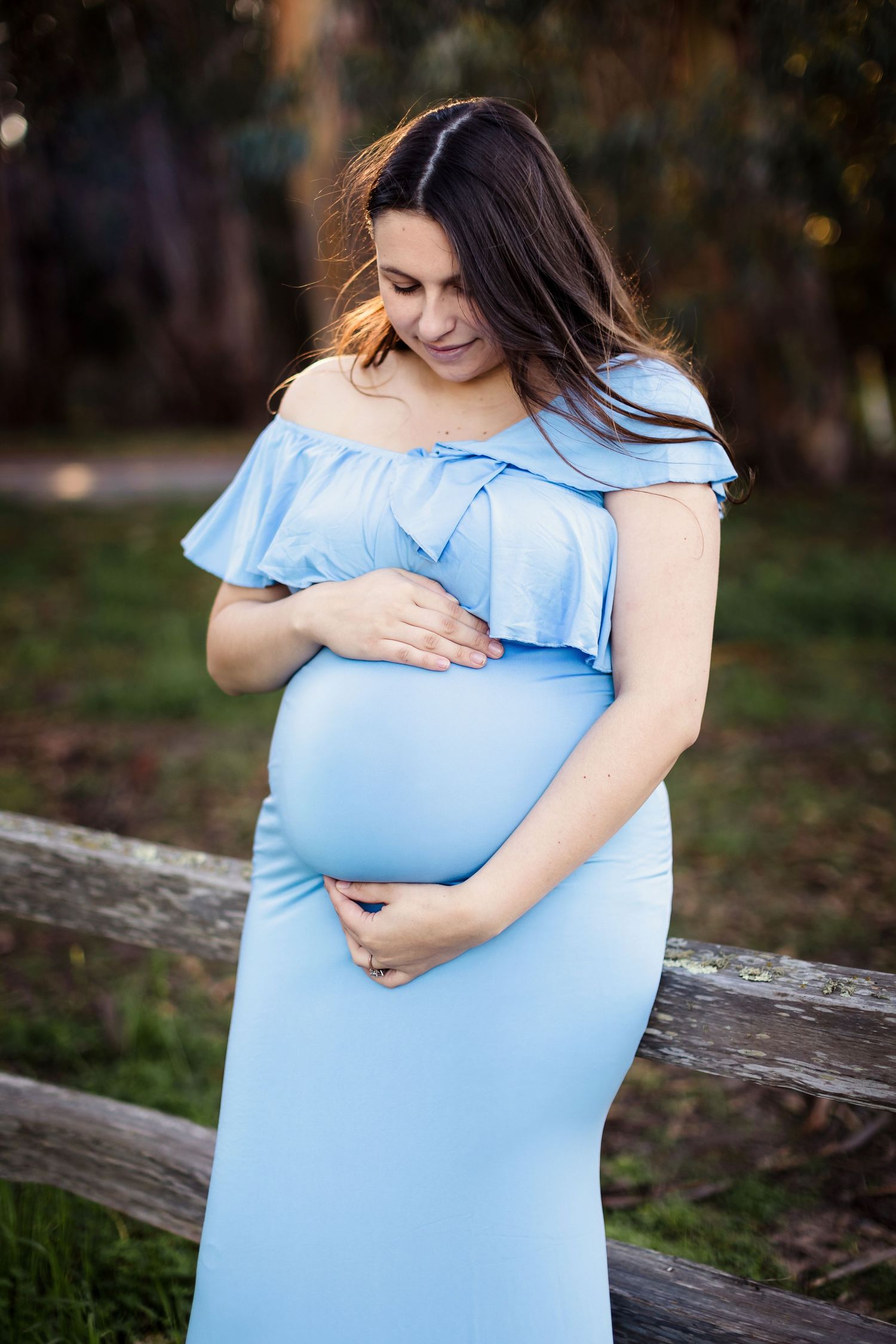 A person in a light blue ruffled maternity dress cradling their baby bump in an outdoor setting at sunset.