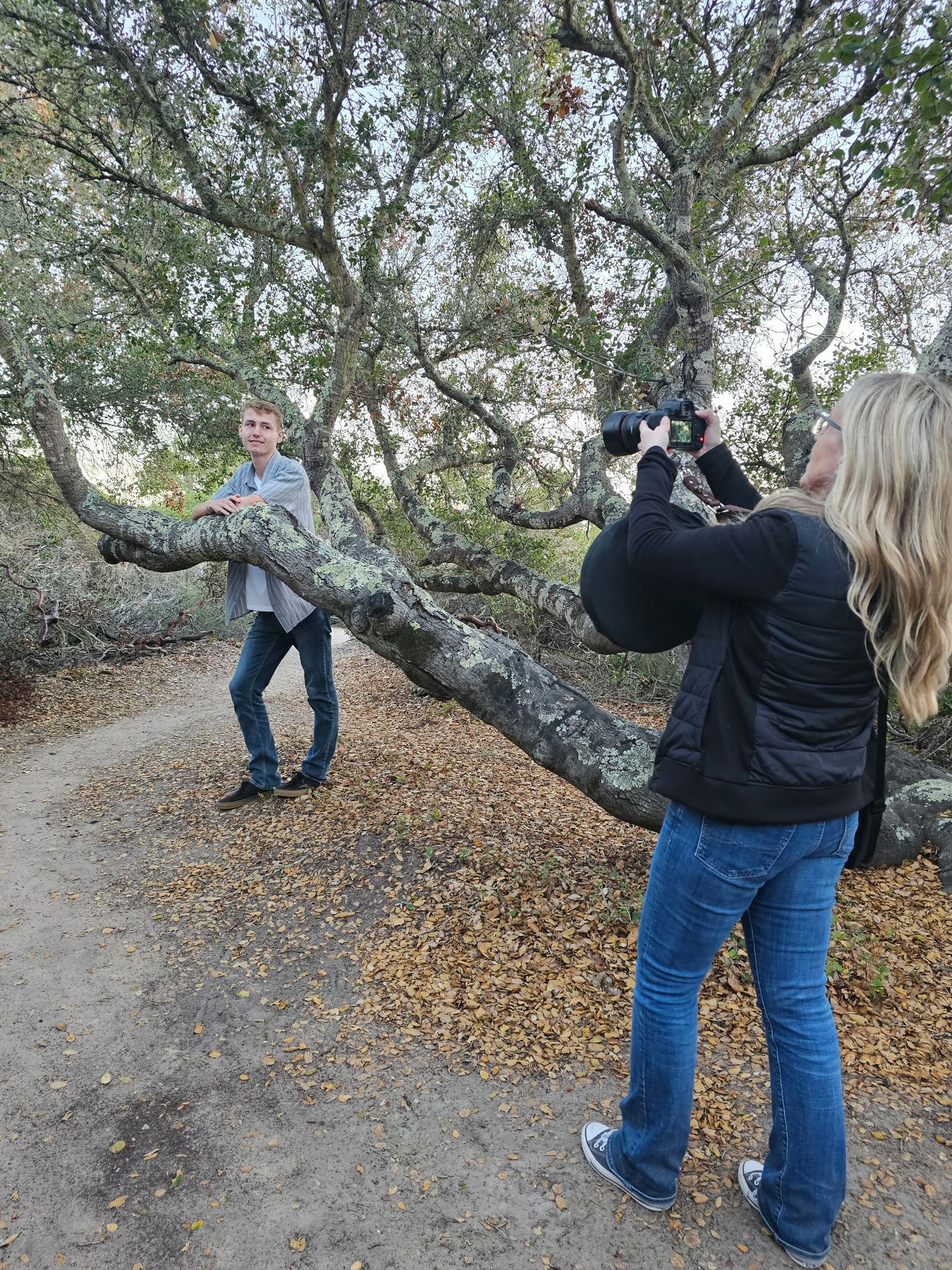 Blonde photographer captures man posing by large twisted oak tree branch on dirt path outdoors.