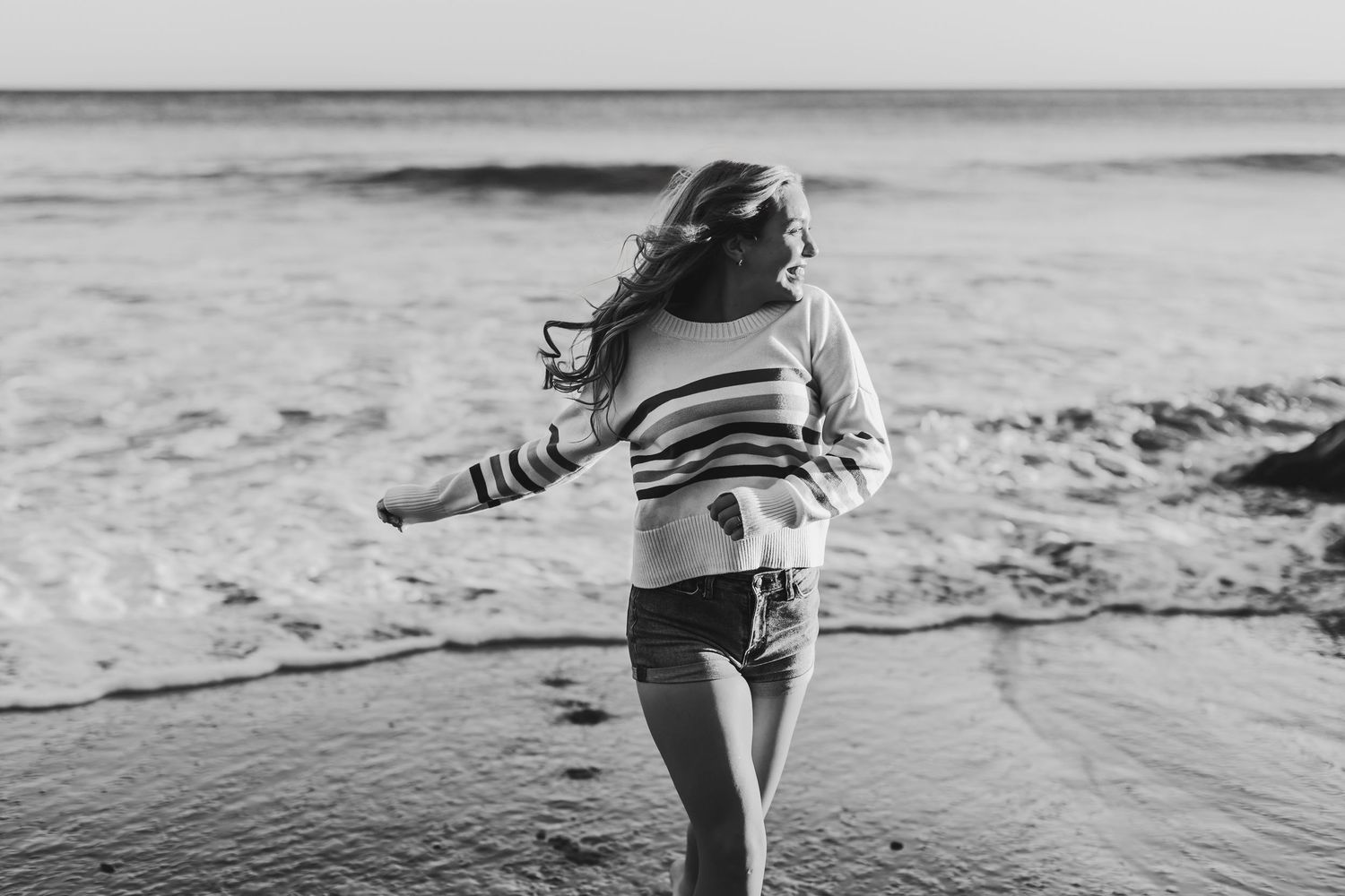 Black and white photo of a woman twirling on a beach with waves behind her, hair blowing in the wind.