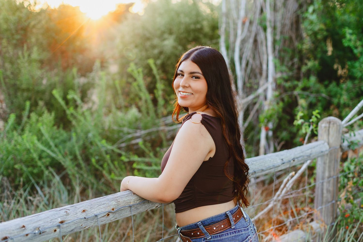 Smiling brunette teen girl in crop top and jeans leaning on wooden fence at golden hour.