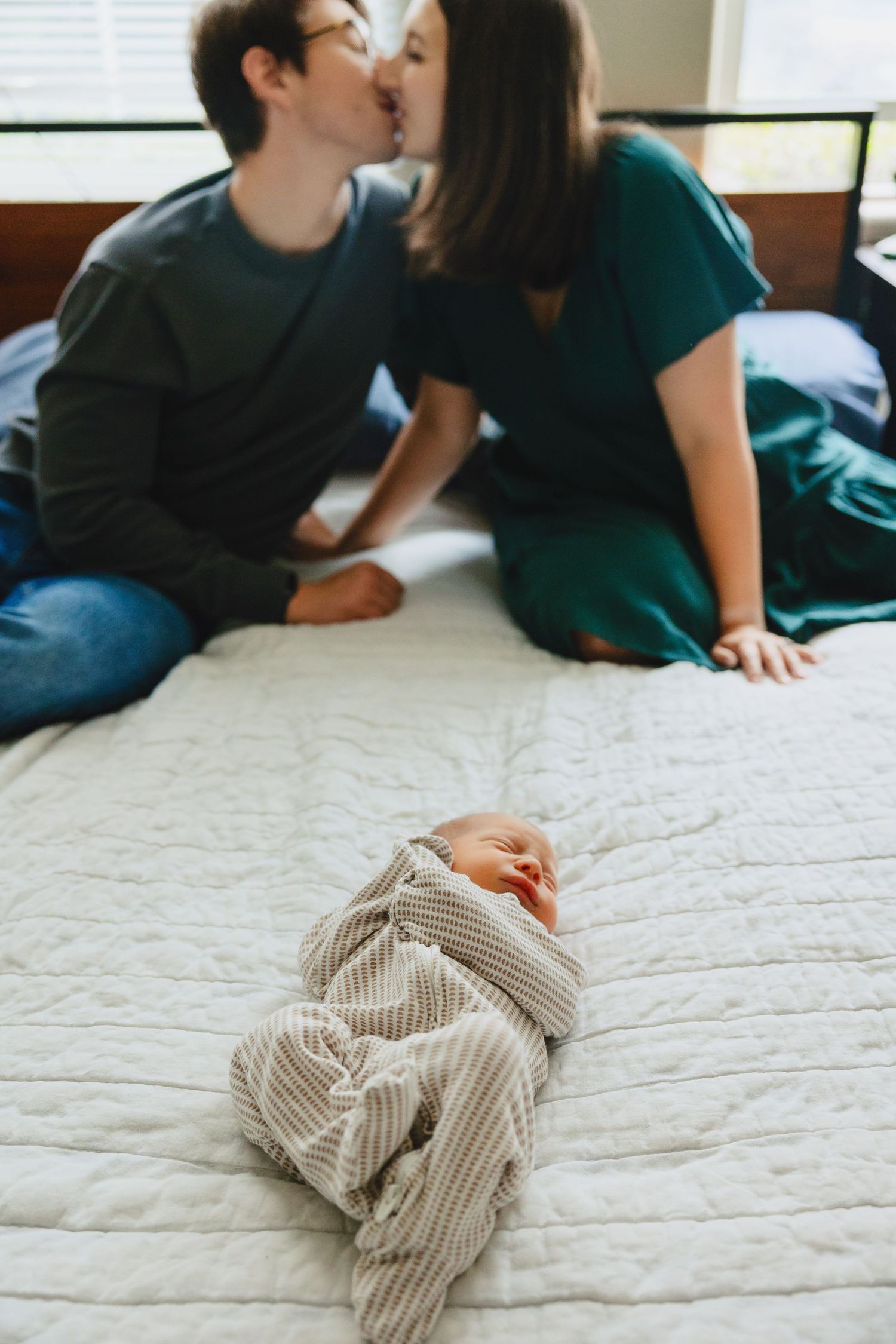 Two people sharing a tender kiss on a bed while their young infant rests peacefully below them on white bedding.