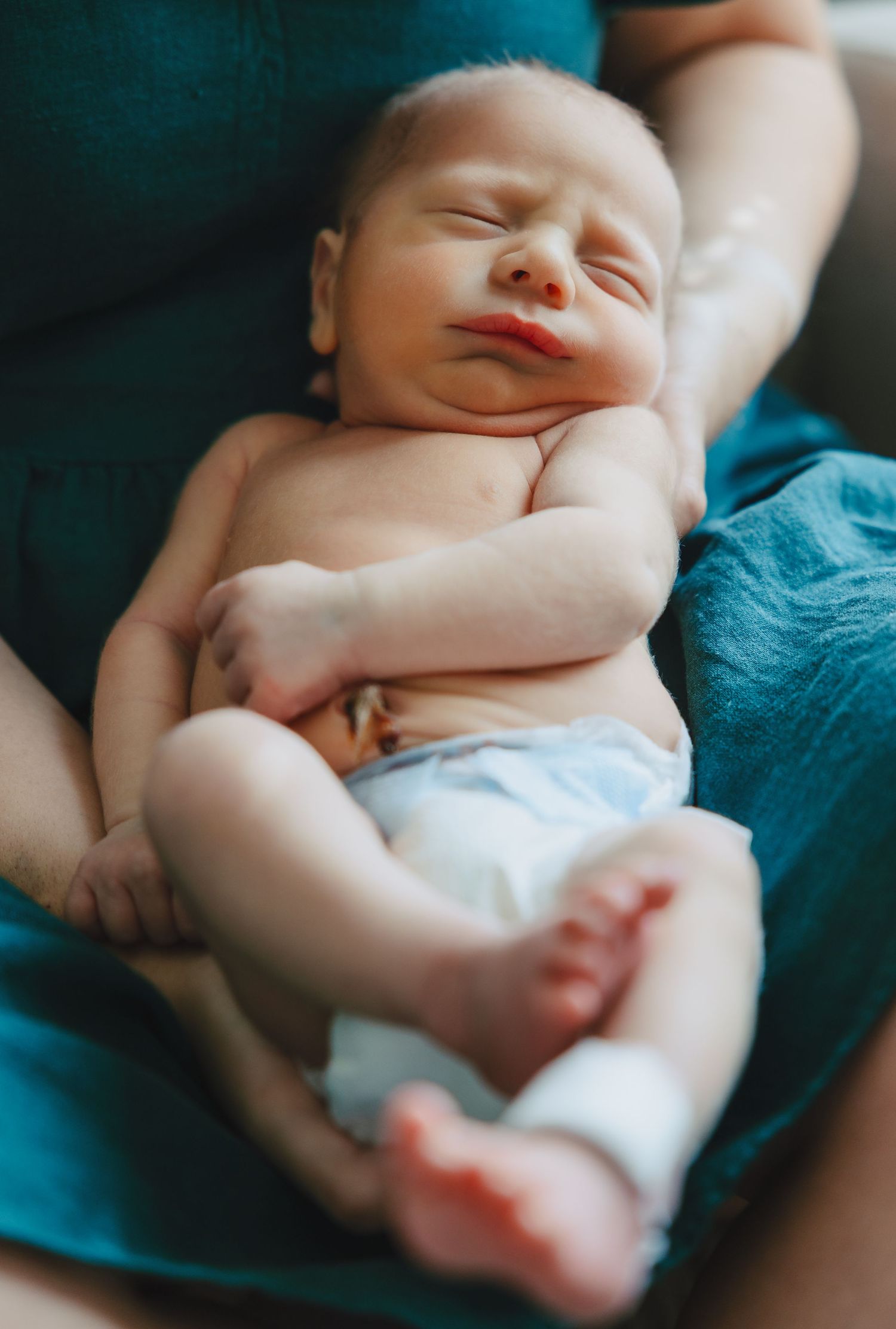 Sleeping newborn baby cradled in mothers arms wearing a teal shirt indoors.