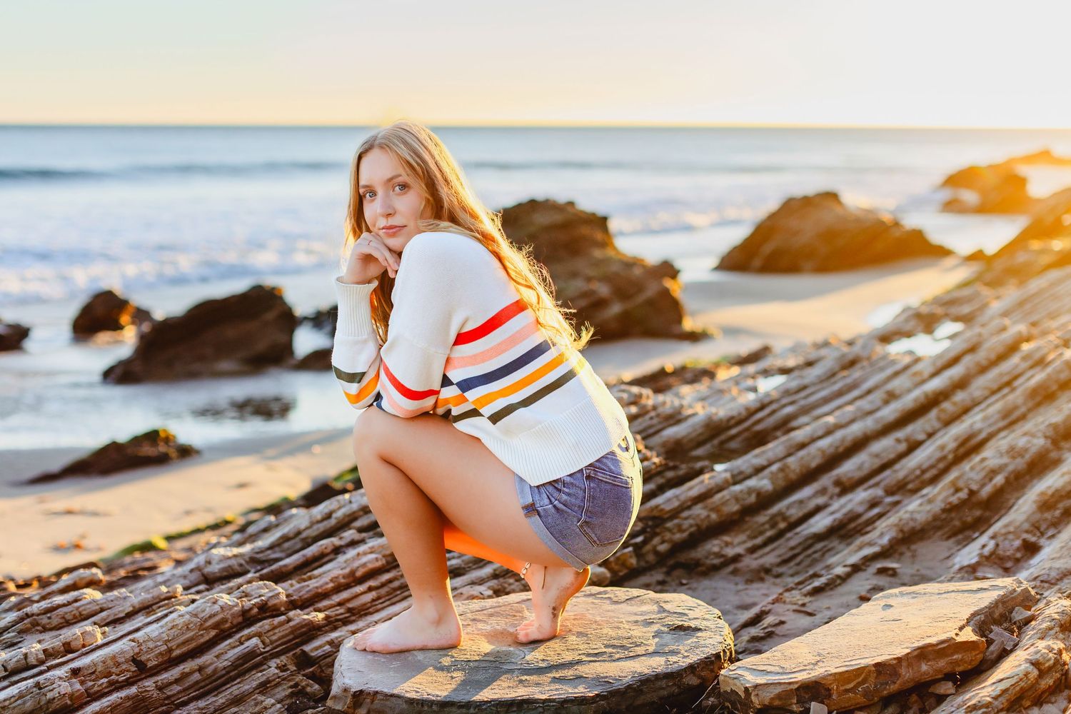 Person in denim shorts and striped sweater crouches on coastal rocks at sunset by the ocean.