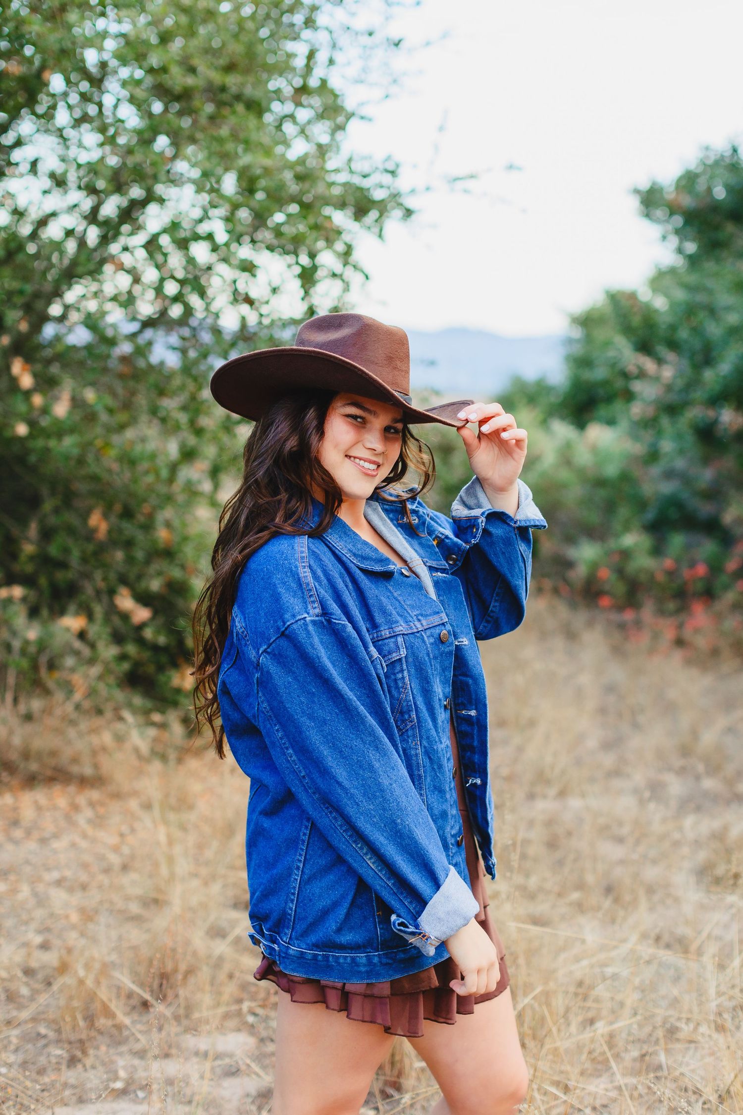 Smiling brunette woman in denim jacket and brown hat poses outdoors in a natural field setting.
