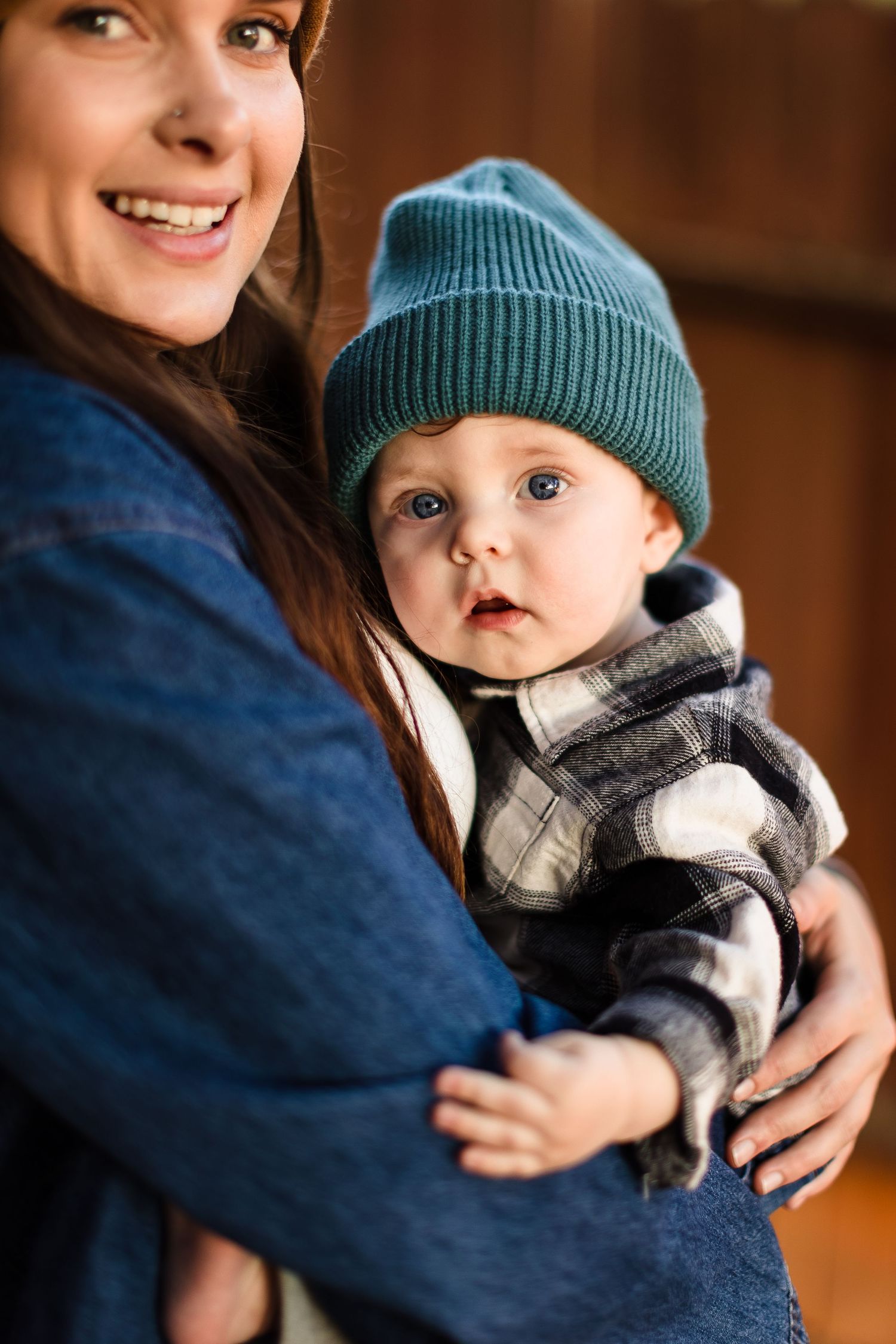 A baby wearing a blue knit beanie and gray sweater is held lovingly while smiling at the camera.