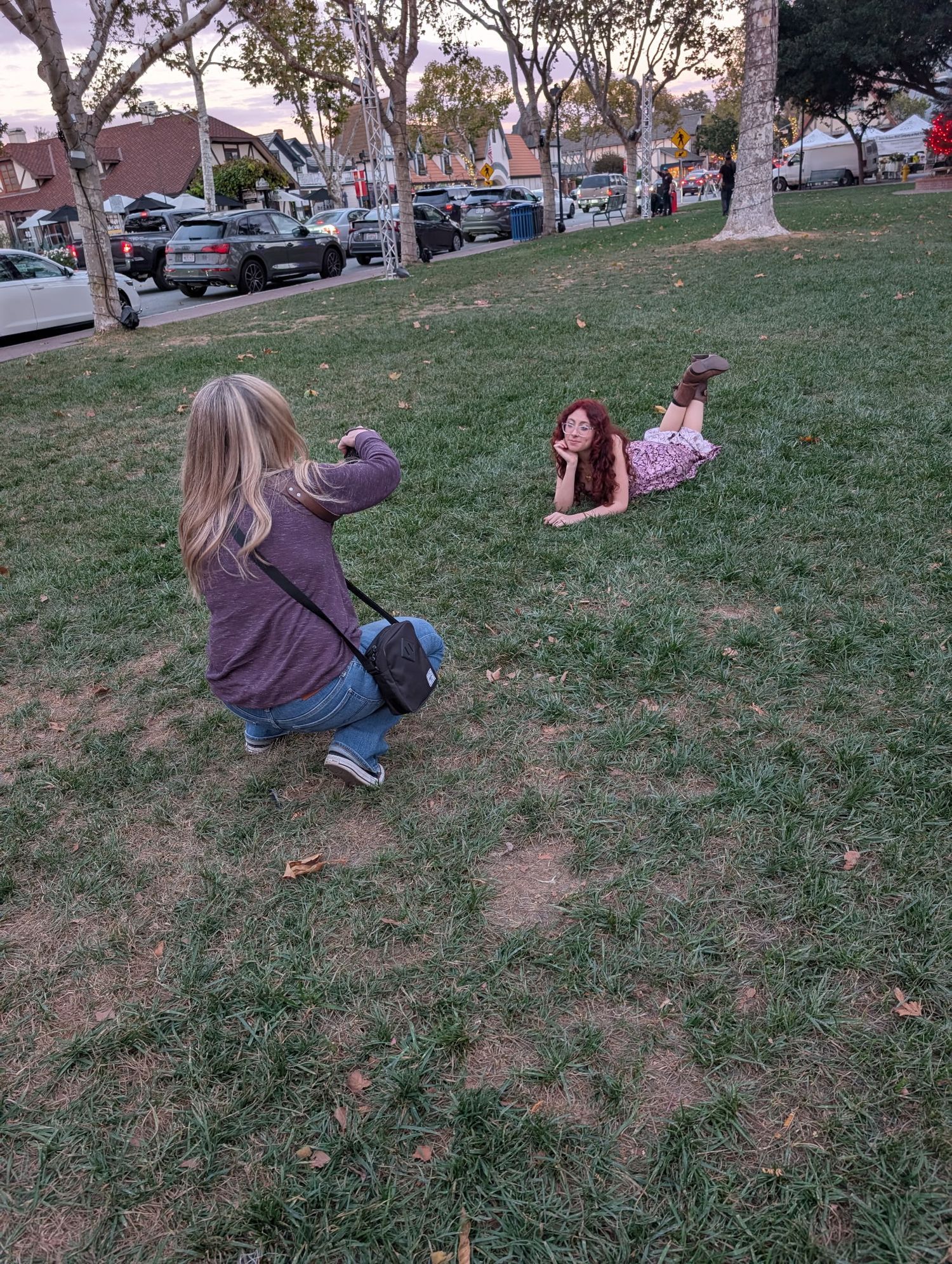 Blonde photographer crouching to photograph red-haired woman posing on green grass in park.