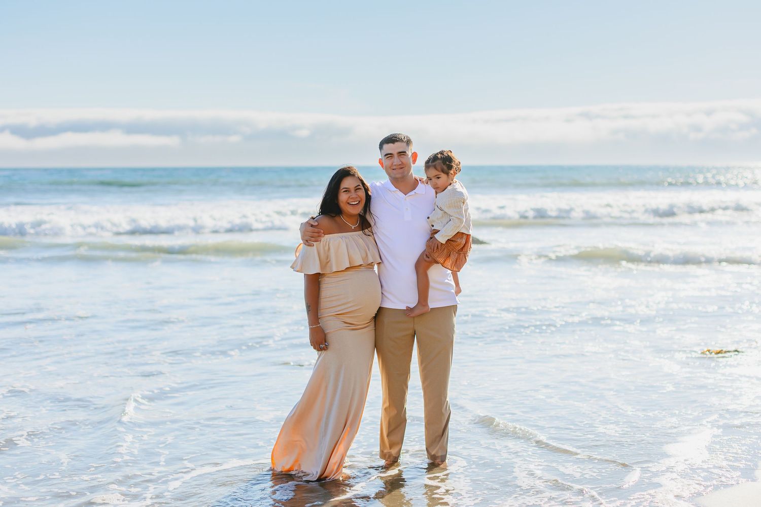 A family stands together on a sunny beach with ocean waves and blue sky in the background.