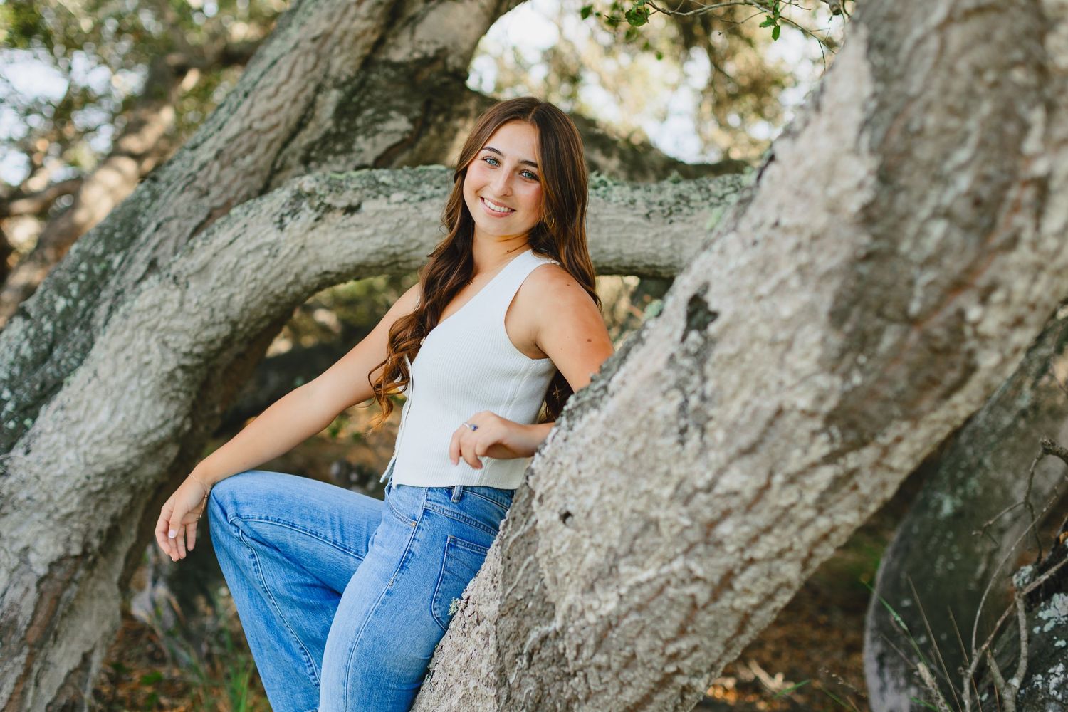 A person in jeans and white top poses casually by large tree trunks on a sunny afternoon.