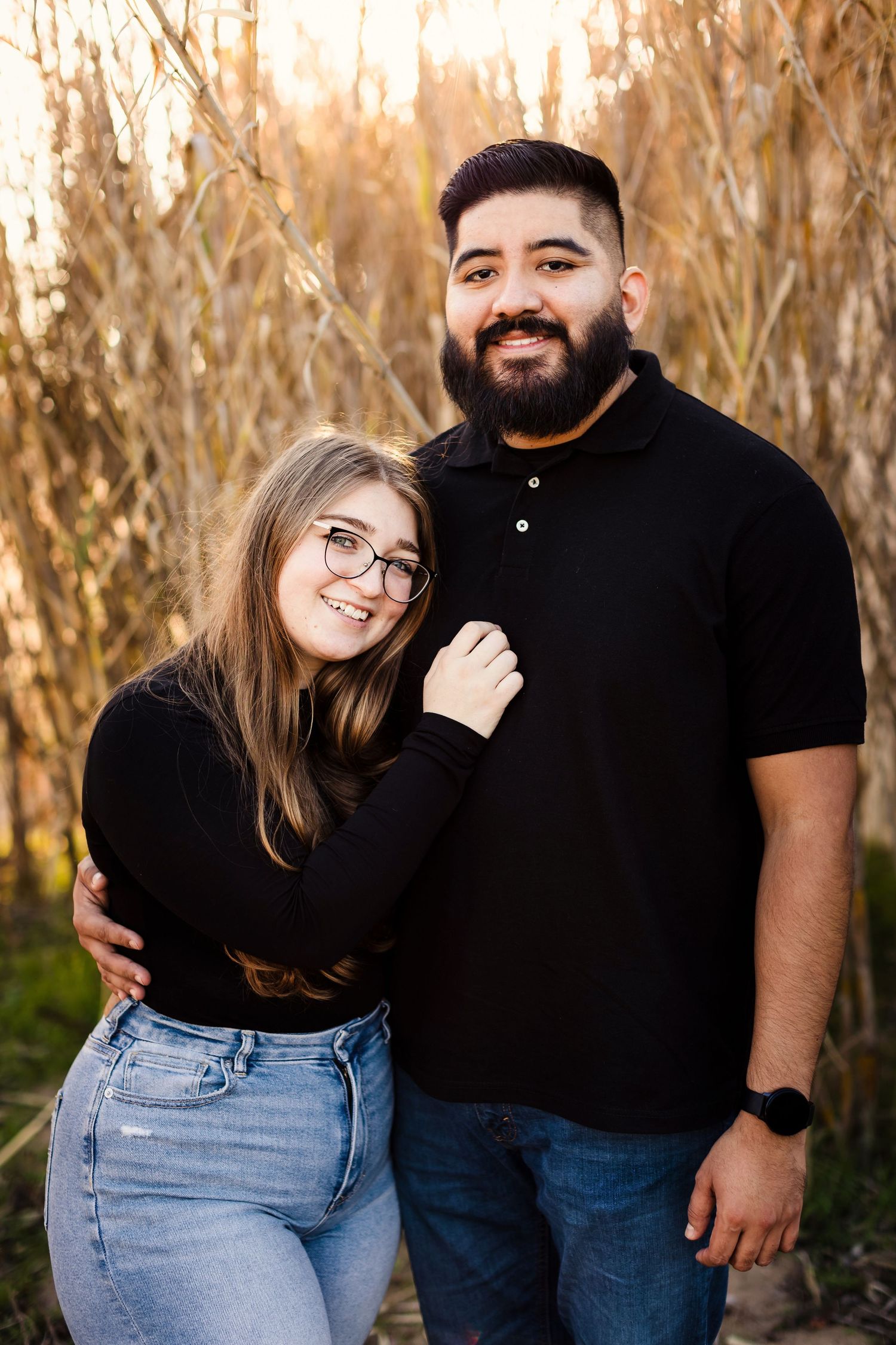 A casually dressed couple embrace in an outdoor setting with tall autumn grass during golden hour lighting.